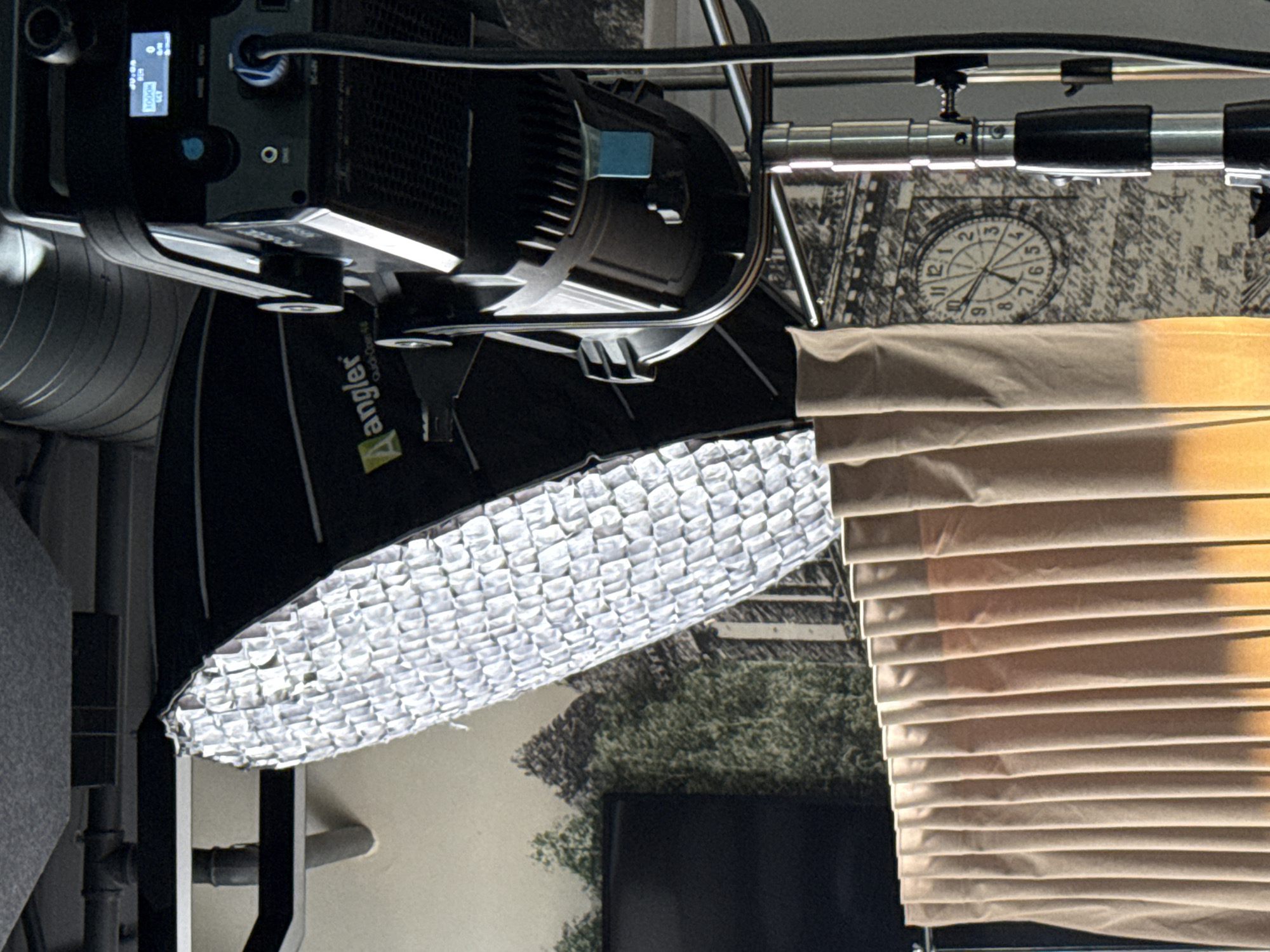 The image depicts a lighting setup, possibly indoors. A light with a softbox, labeled "Aangler", is prominent. The background features a clock face and a nature scene, partly obscured by closed window blinds. The blinds appear to be tan or beige. The overall impression is of a studio or a room being used for photography or videography. There is no clear evidence to confirm the location being Yerevan, Armenia.