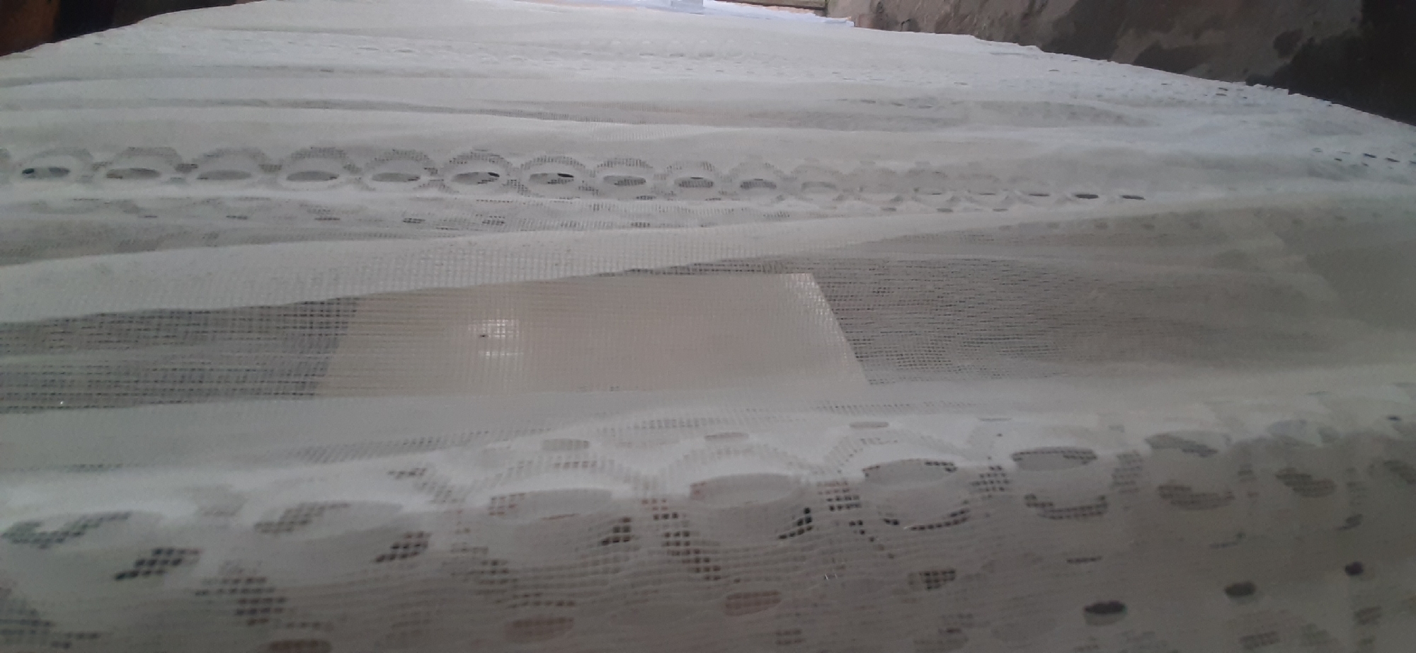 A white lace curtain is prominently featured in the foreground, obscuring much of the scene. The curtain has a textured appearance with intricate patterns of holes and decorative borders. Behind the curtain, a rectangular, pale object is visible, possibly a piece of furniture or a wall. The background on the right side of the image shows a dark, textured surface, likely a floor or ground. The overall lighting suggests an indoor environment. There are no people or animals visible in the image. The location in Yessentukskaya, Russia, is not discernible from the visual information provided in the image.