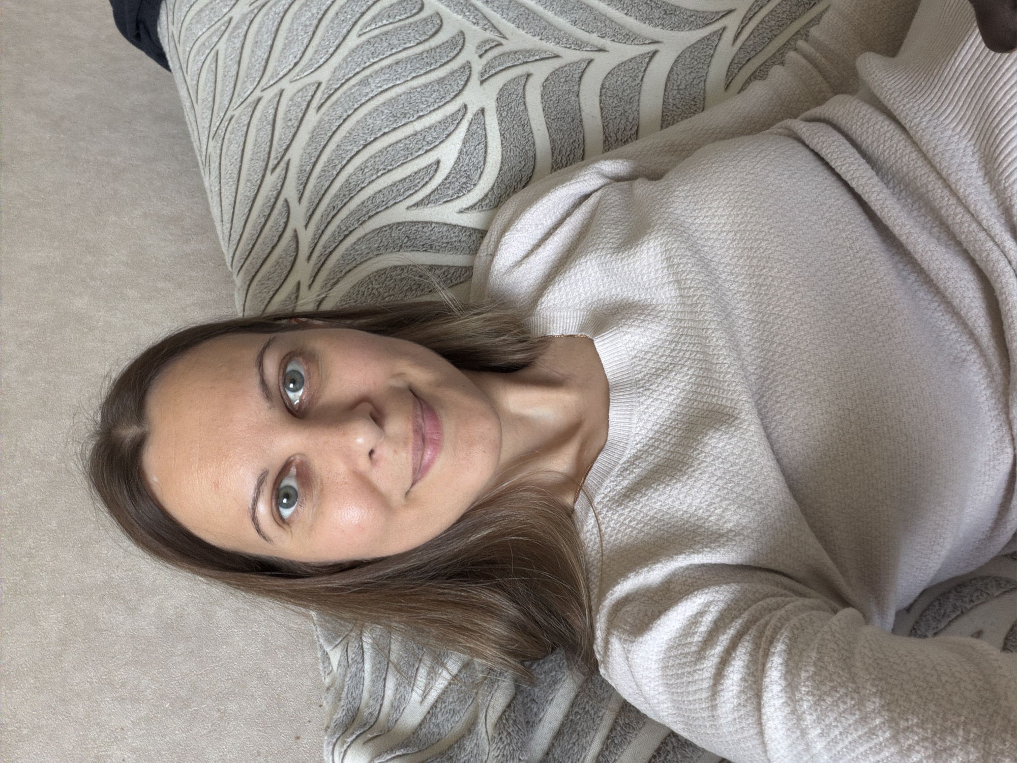 A woman is pictured sitting on a sofa in a room, facing the camera and smiling. She has brown hair that falls past her shoulders and blue-green eyes. She is wearing a light-colored, long-sleeved sweater. The sofa has a patterned fabric with alternating stripes and leaf-like designs in gray and white. The background appears to be a wall with a textured wallpaper or paint finish in a neutral tone. There are no visible identifiers of a specific location in Izhevsk, Russia.