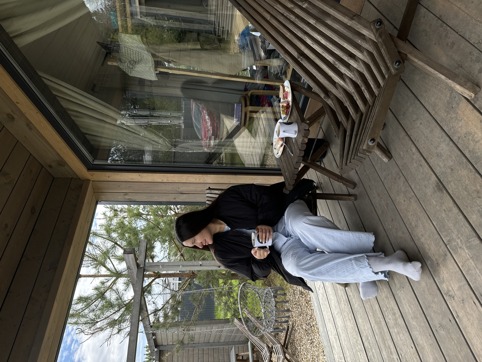 The image shows a woman sitting on a wooden deck in Литвиново, Russia. She is wearing a black jacket and light blue pants, and she is holding a white mug. A small wooden table with a plate of food and a cup sits next to her. There is a large window behind her, reflecting the interior of the building and a red car outside. The deck is made of wooden planks, and there is a slatted wooden structure nearby. Outside, a tree and a portion of a swing set are visible.