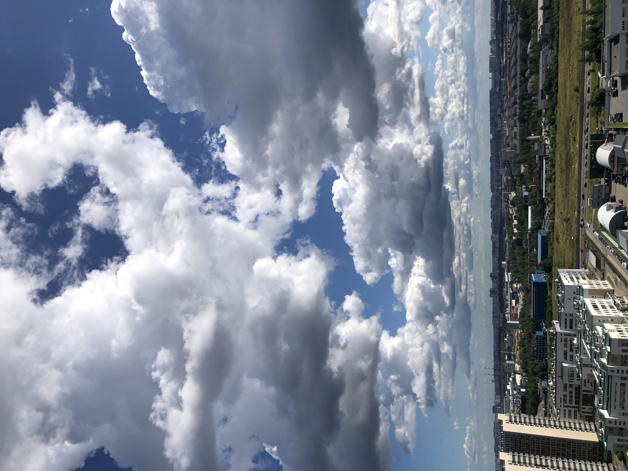 The image shows a cityscape with a vast expanse of sky filled with dramatic clouds. In the foreground, several modern residential buildings are visible, characterized by their white facades and numerous windows. The buildings are arranged in a dense urban layout. Beyond the residential areas, the landscape transitions into industrial zones with various factory buildings, warehouses, and what appear to be construction sites with cranes. A highway runs through the midground, with a steady stream of cars. Further in the distance, a sprawling city skyline stretches across the horizon, with tall buildings and industrial structures. The sky is a striking contrast of deep blue patches and voluminous white and gray clouds, suggesting a dynamic weather pattern. The overall scene depicts a developed urban and industrial environment under a partly cloudy sky.