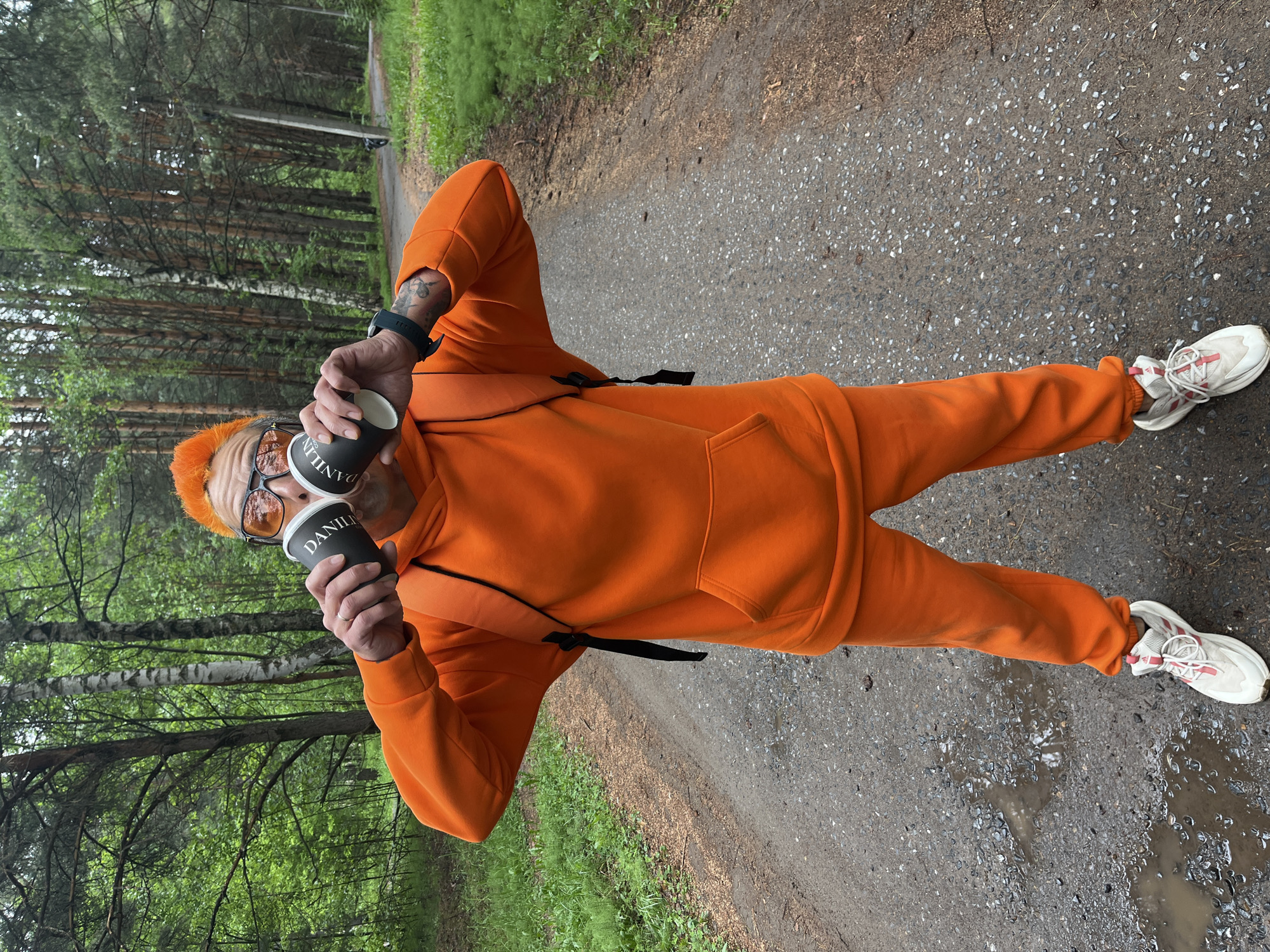 A man in an orange tracksuit stands on a paved path, holding two black coffee cups to his ears like headphones. He has orange hair and sunglasses. The path is damp, with puddles and a scattering of small stones. On either side of the path are trees. The coffee cups are labeled "Danilin." He wears white running shoes and a wristwatch. The scene is in Chaikovsky, Russia.