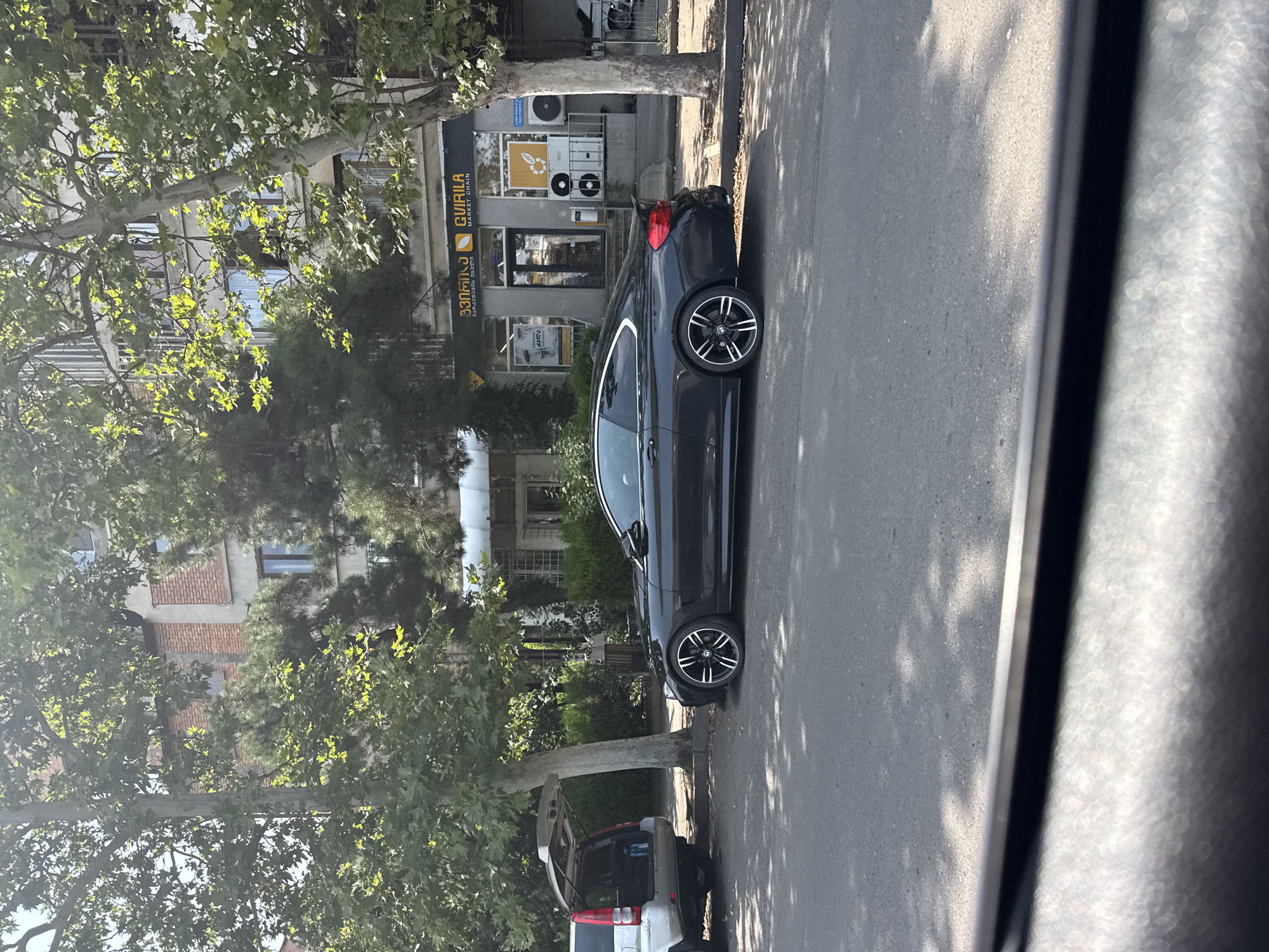 In Tbilisi, Georgia, on a sunny day, a dark gray BMW 4 Series coupe is parked on a paved street alongside a sidewalk. The car is positioned facing right in the frame. To the left of the BMW, a white SUV with its rear hatch open is parked. Behind the vehicles, a building with multiple stories, featuring brick and concrete facades, is visible. Trees with green foliage line the street, casting shadows on the pavement. A store with the name "GVIRILA MARKET CHAIN" is situated on the ground floor of the building, identified by its yellow and black signage. The scene appears to be in an urban residential or commercial area.