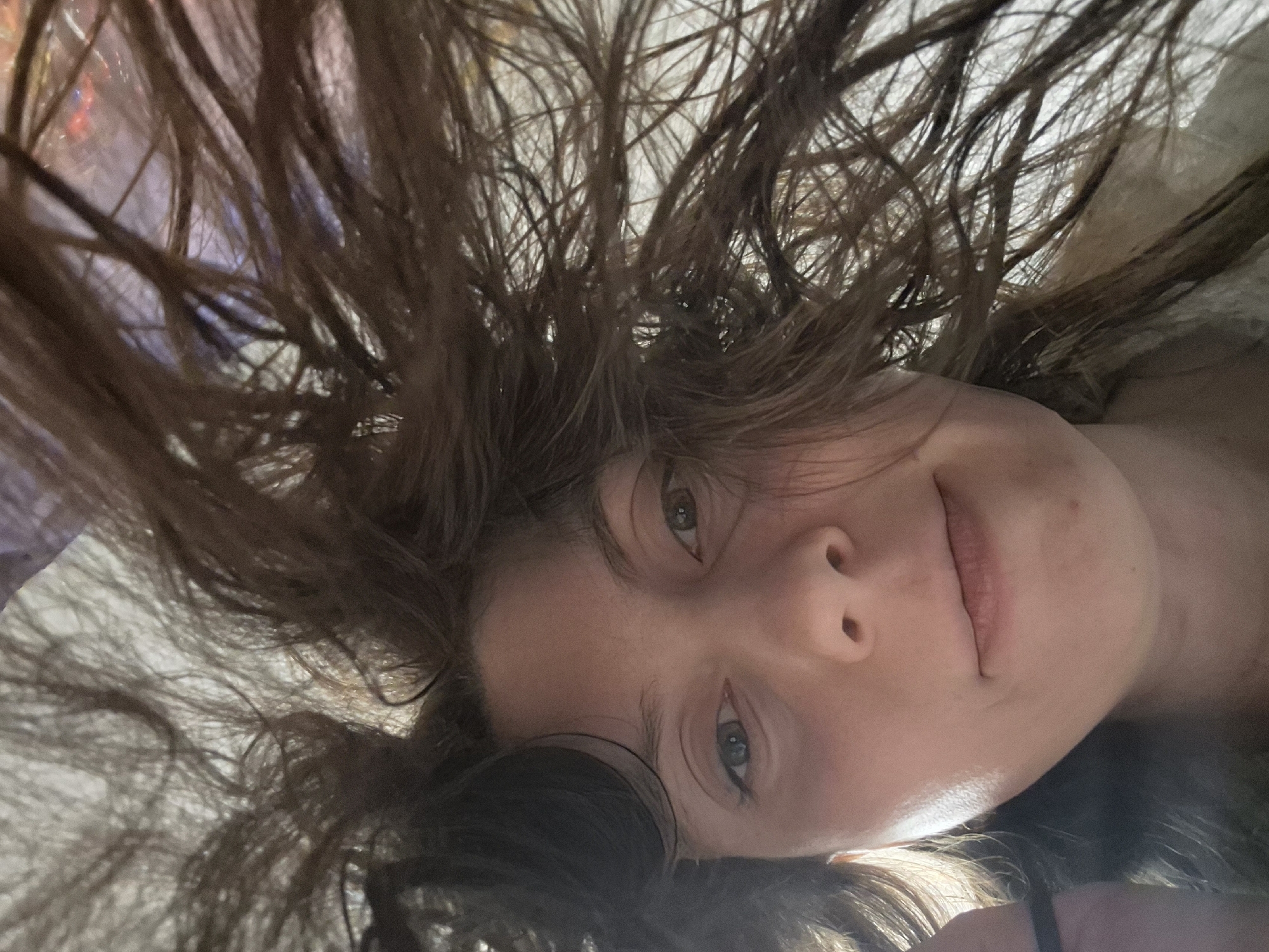 A close-up, slightly skewed, selfie-style photograph shows a person with light skin and brown hair spread around their face. The person's eyes are visible, and they appear to be looking directly into the camera. The background is a mix of indistinct light and dark areas. The location is Unknown, Unknown. There is no other information about the city location to identify.