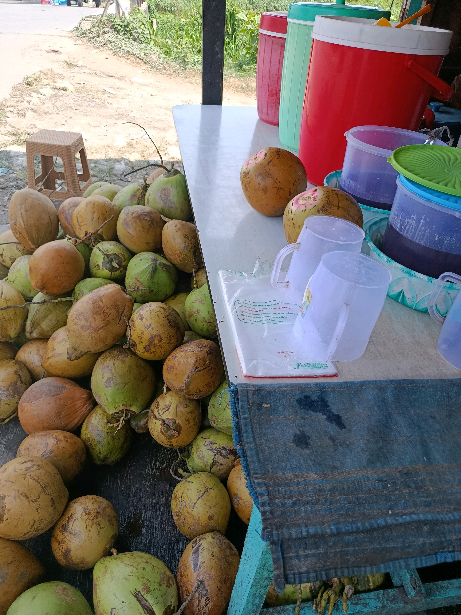 The image depicts an outdoor coconut stand in Babelan, Indonesia. A large pile of fresh coconuts, a mix of green and brown, is displayed on a dark surface in the foreground. To the right, on a white table, are several clear plastic cups and a clear plastic bag with printed text. Behind these, a large red cooler is visible, along with a green and a red thermos. The background shows a dusty road, green vegetation, and a glimpse of what appears to be a street. The lighting suggests it is daytime.