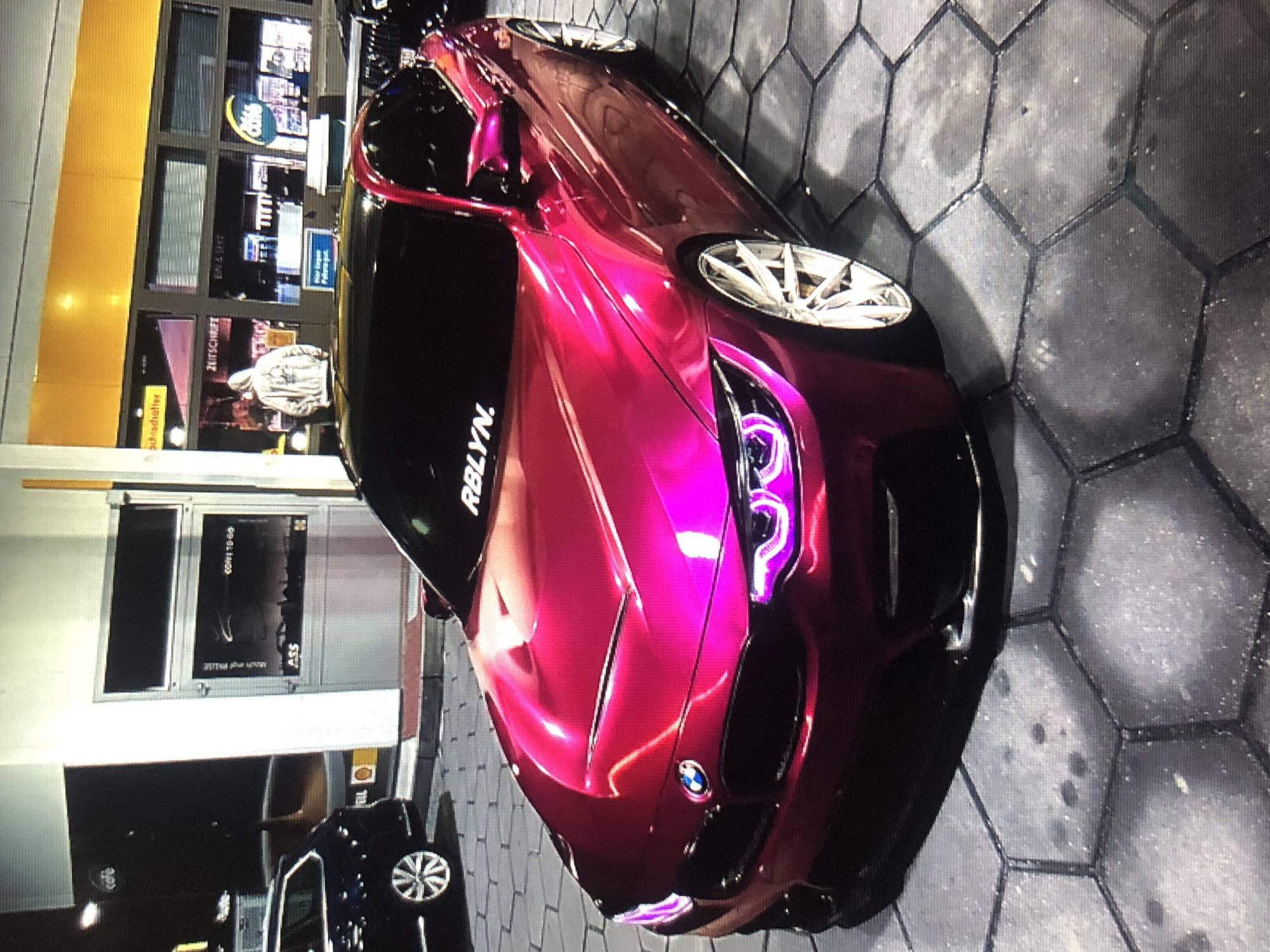 A maroon BMW M4 with custom headlights is parked in front of a Shell gas station in an urban setting. The car has the word "RBLYN" on its windshield and custom wheels. The flooring appears to be hexagonal pavers. A black sedan with silver rims is parked to the side, partially visible. The gas station has large windows, with signage and advertisements visible inside. The photo is taken at night or in low-light conditions. The precise location within Lhokseumawe cannot be determined from the image.