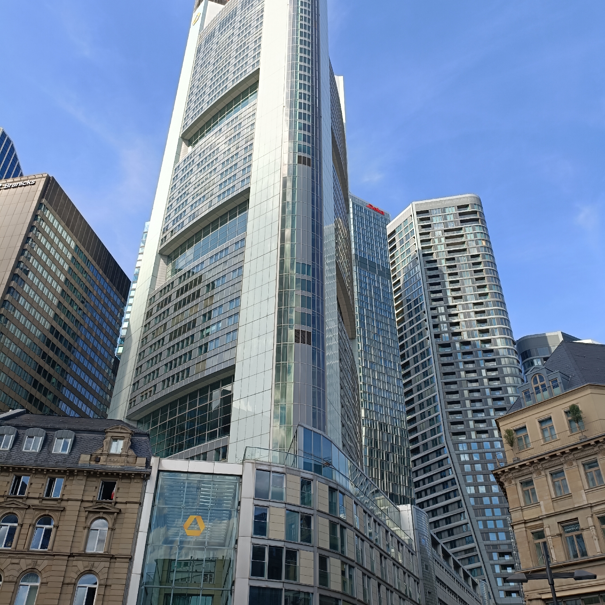 The image shows a low-angle view of several modern skyscrapers and an older building in Frankfurt, Germany. The most prominent building is a very tall, white skyscraper with a glass facade, reflecting the blue sky and clouds. To its right, another tall, grey skyscraper has a distinctive curved facade with many windows. In the foreground, on the left, an older, beige building with ornate architectural details and mansard roof windows is visible. A modern glass structure with a yellow triangular logo is attached to the side of this older building. The sky is clear and blue. The overall scene depicts an urban environment with a mix of architectural styles.