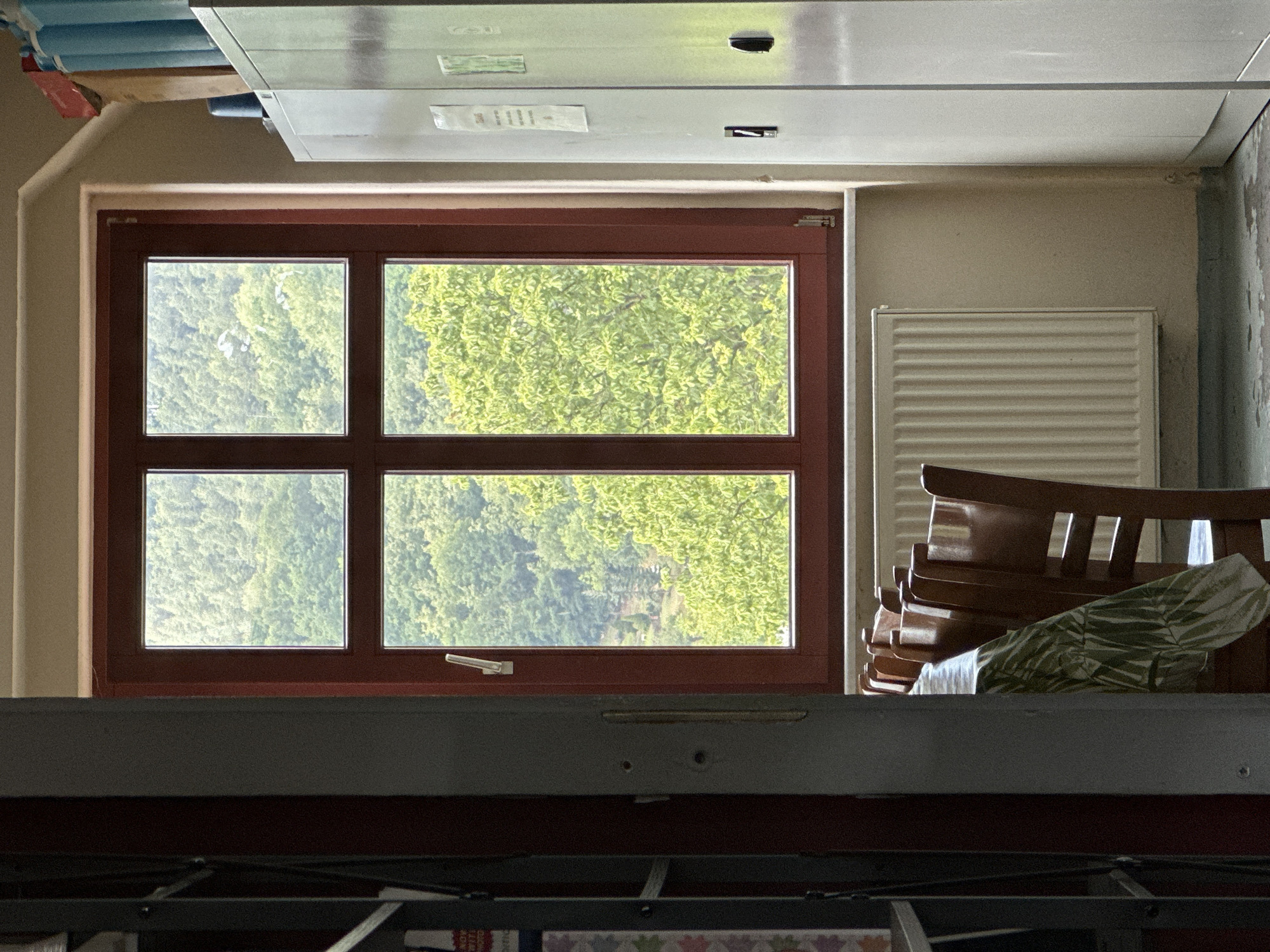 The image depicts an interior scene from what appears to be a room with a large window overlooking a forest. The room contains a table covered with a patterned tablecloth and several wooden chairs. To the right of the window is a tall, white metal cabinet, and to the left, a bookshelf is partially visible. A radiator is located beneath the window. The view through the window reveals a dense canopy of green trees, suggesting a natural, possibly rural or park-like setting outside. The presence of a table and chairs suggests a communal area, possibly a break room or dining space. The overall atmosphere is one of quiet occupancy, with no people present in the frame. The scene is well-lit, with natural light streaming in from the window. Given the context provided, this room is located in Bad Blankenburg, Germany.