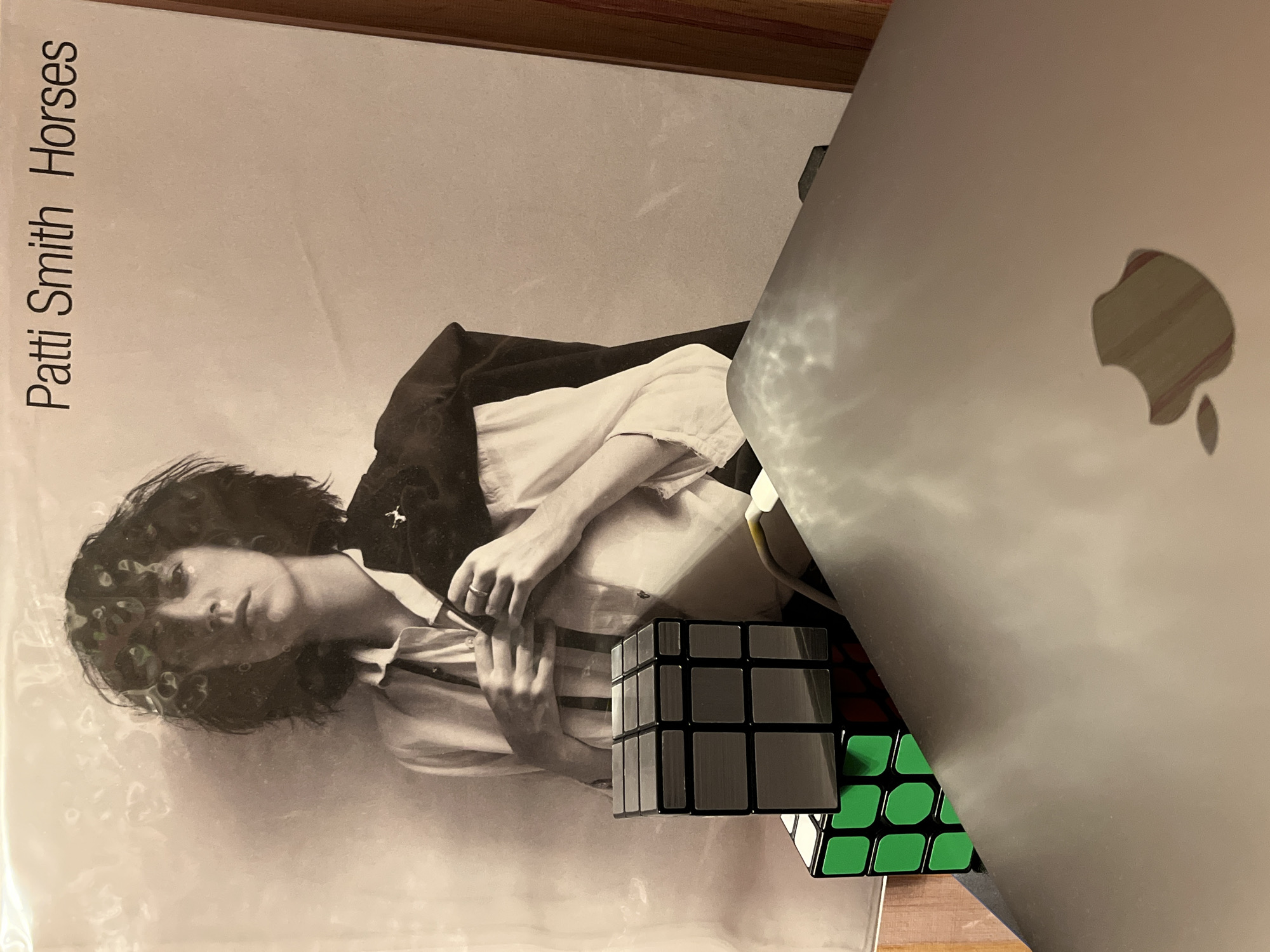 A black and white album cover for "Patti Smith Horses" is displayed. In the foreground, a silver laptop with an Apple logo is partially visible. To the left of the laptop, two Rubik's cubes are stacked. The top cube is silver and black, while the bottom cube has green sides. The background features a portrait of Patti Smith, who is wearing a white shirt and a dark jacket draped over her shoulder. Her hands are adjusting the buttons of her shirt. The image appears to be taken indoors, possibly in a residential setting, with no discernible city or location information.