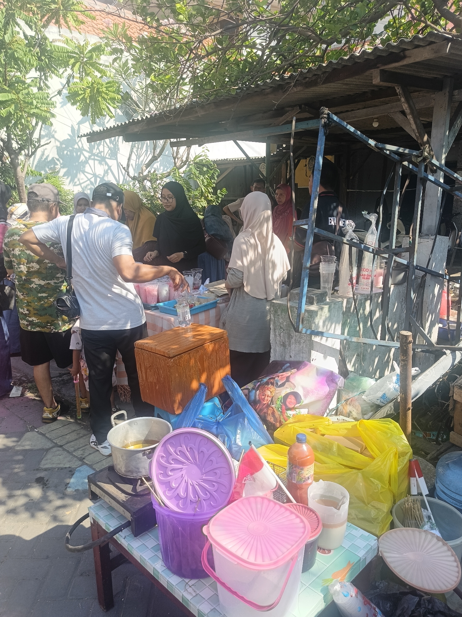 In Gedangan, Indonesia, a street food stall is depicted under a makeshift corrugated metal roof, offering refreshments. The scene is bustling with activity as several individuals, some wearing hijabs, engage with the vendor or purchase items. In the foreground, a variety of containers are visible, including purple and pink plastic buckets, a large aluminum pot on a portable stove, and plastic cups. Yellow and blue plastic bags are piled up, some containing food items and others appearing to be packaging. A bottle of orange liquid and skewers for food preparation are also present. The ground is paved with asphalt, showing wear and tear. The overall impression is of a lively, informal market setting on a sunny day.
