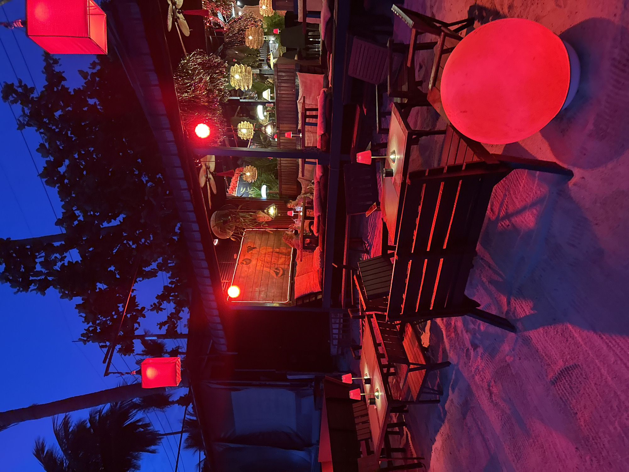 The image depicts an outdoor seating area at dusk, likely a restaurant or bar, located in the Ko Pha-ngan District of Thailand. The scene is illuminated by red and pink ambient lighting, casting a warm glow on the sandy ground and wooden furniture. Several tables and chairs are arranged on the sand, with some tables featuring small red table lamps. In the background, there is a covered seating area with more seating, hanging lanterns, and ceiling fans. Palm trees and dense foliage are visible against the dark blue twilight sky. A large, illuminated red orb sits on the sand in the foreground, adding to the vibrant atmosphere. Two individuals are seated in the background seating area, partially obscured by the lighting and distance.