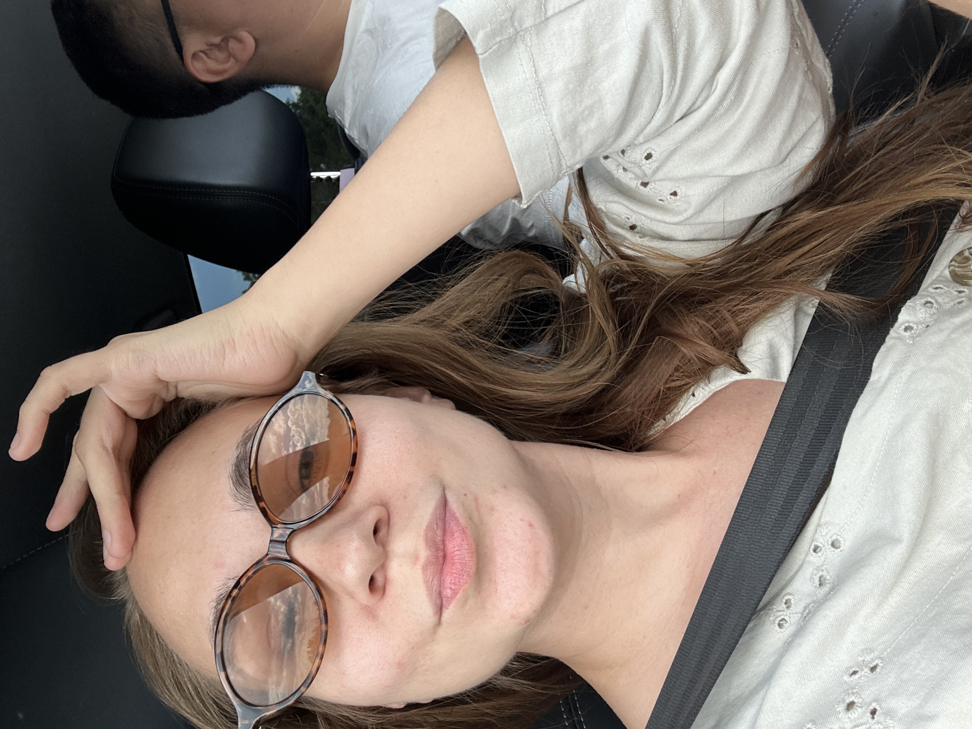 A woman with auburn hair is in the passenger seat of a car, looking at the camera. She is wearing a light-colored top with intricate cutwork detailing around the neckline and sleeves, and a dark seatbelt across her chest. Large, tortoiseshell-patterned sunglasses with amber lenses obscure her eyes, and her left hand is raised to her forehead. The reflections in her sunglasses show a sky with clouds. To her right, a man with short dark hair and glasses is visible in the driver's seat. The interior of the car appears to be dark, with a black leather headrest visible behind the woman. The overall scene suggests a casual journey, possibly during daylight hours given the lighting and reflections. The image does not provide specific visual cues to indicate a location within the city of Pervomaiskoe, Russia.
