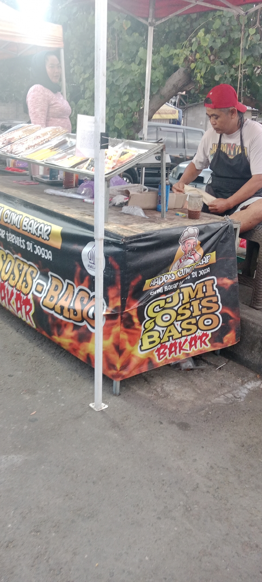 A street food vendor in Ngemplak, Indonesia, is captured in a medium shot. The vendor, a man wearing a red baseball cap and a black apron over a light-colored t-shirt, is seated behind a food cart. He is shown in the process of preparing food, holding a piece of dough or wrapper in his hands. A glass of dark liquid sits on the counter in front of him.
The food cart itself is adorned with a large black banner featuring colorful text and graphics of flames. The text appears to advertise food items like "CUMI BAKAR" (grilled squid), "SOSIS" (sausage), and "BASO BAKAR" (grilled meatballs). A cartoon illustration of a chef is also part of the banner's design.
In the background, a woman in a pink and white patterned shirt and a dark headscarf stands behind the counter, looking towards the left of the frame. Several trays of food are displayed on the cart, including skewers of what appear to be grilled meat or seafood, and other food items. The scene is outdoors, with greenery and trees visible behind the stall. The ground is paved, suggesting a street or roadside location. A white support pole for the stall's awning is prominent in the foreground.