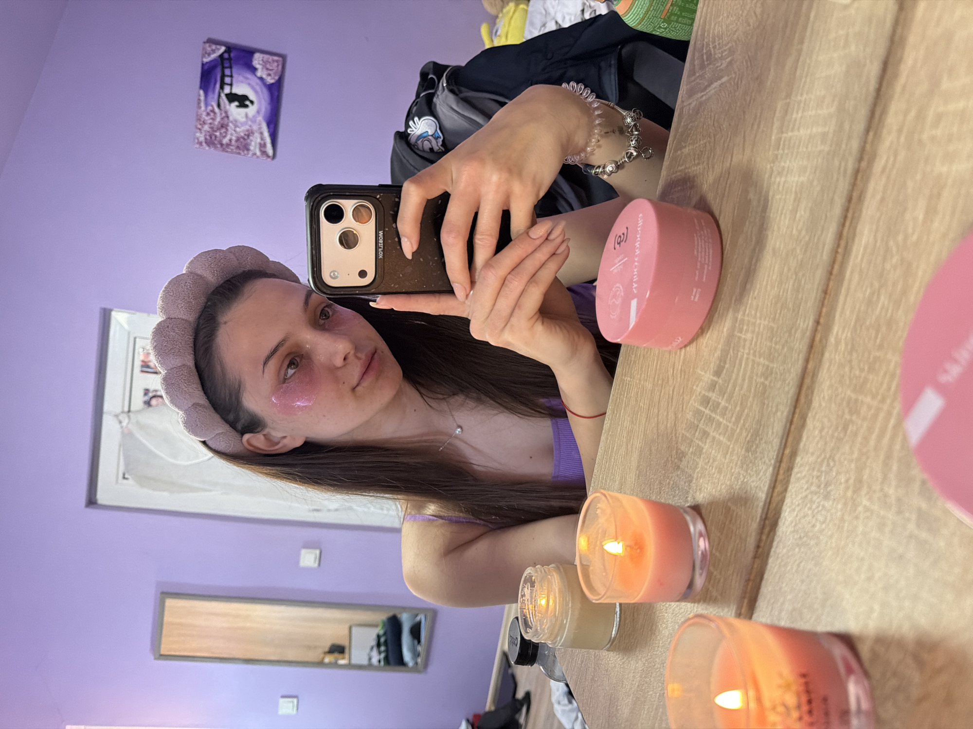 A young woman is taking a selfie in a room with light purple walls. She is wearing a soft, padded headband and pink eye patches under her eyes. She holds a black smartphone in her right hand to capture the image. Her long, brown hair falls over her left shoulder.
On the right side of the frame, a wooden surface (likely a tabletop or vanity) is visible, reflecting the woman and objects on it. There are several lit candles in glass containers with various colors (yellow and pink) and pink cosmetic jars. A bracelet adorns her wrist.
In the background, a framed print hangs on the wall. Below it is a framed mirror. Clothes and other items are scattered on the floor.