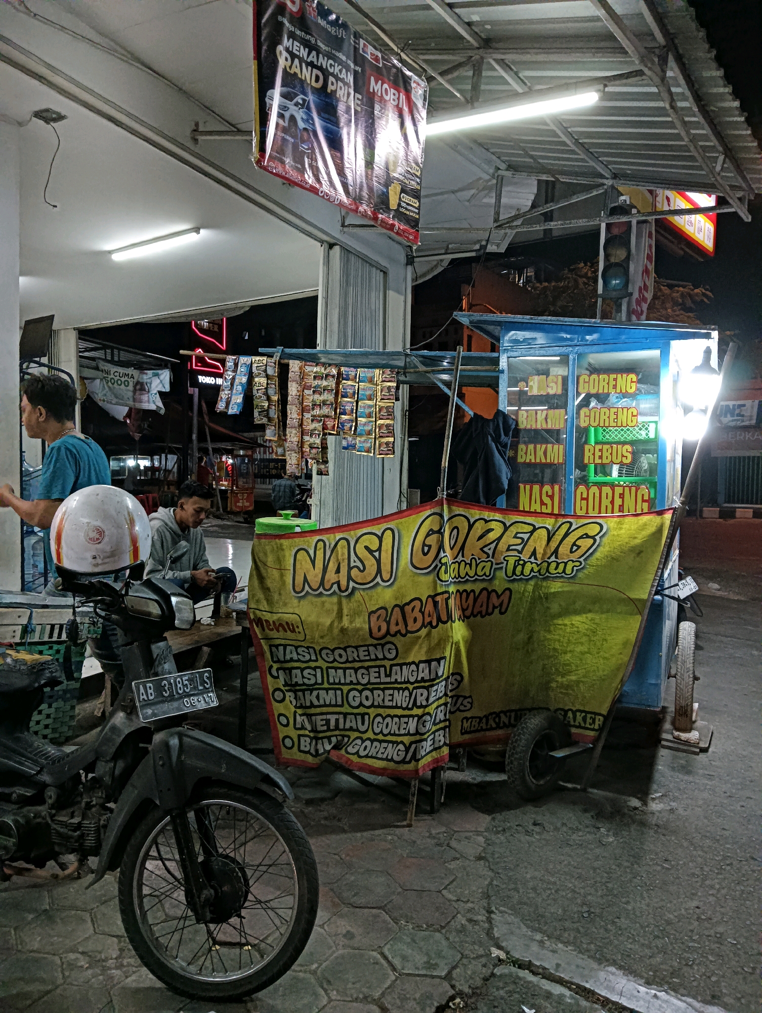 This image captures a nighttime scene in Purworejo, Indonesia, focusing on a mobile food stall selling "Nasi Goreng Jawa Timur" (East Javanese Fried Rice). The stall is a blue cart with a yellow banner displaying the menu, which includes various fried rice and noodle dishes. Inside the cart, illuminated by lights, are stacks of what appear to be containers or ingredients. Adjacent to the cart, a motorcycle is parked, equipped with a white helmet on its handlebars. Two men are present: one in a blue shirt stands to the left, and another in a gray hoodie sits on a wooden crate to the right, looking at his phone. The environment suggests a street-side location, with a paved ground and overhead structures, possibly part of a shop or covered market. A prominent banner hanging above the stall advertises a "GRAND PRIZE" with images of cars, suggesting a promotional event. The overall lighting is artificial, coming from the stall and overhead lamps, creating a slightly dark and atmospheric setting.