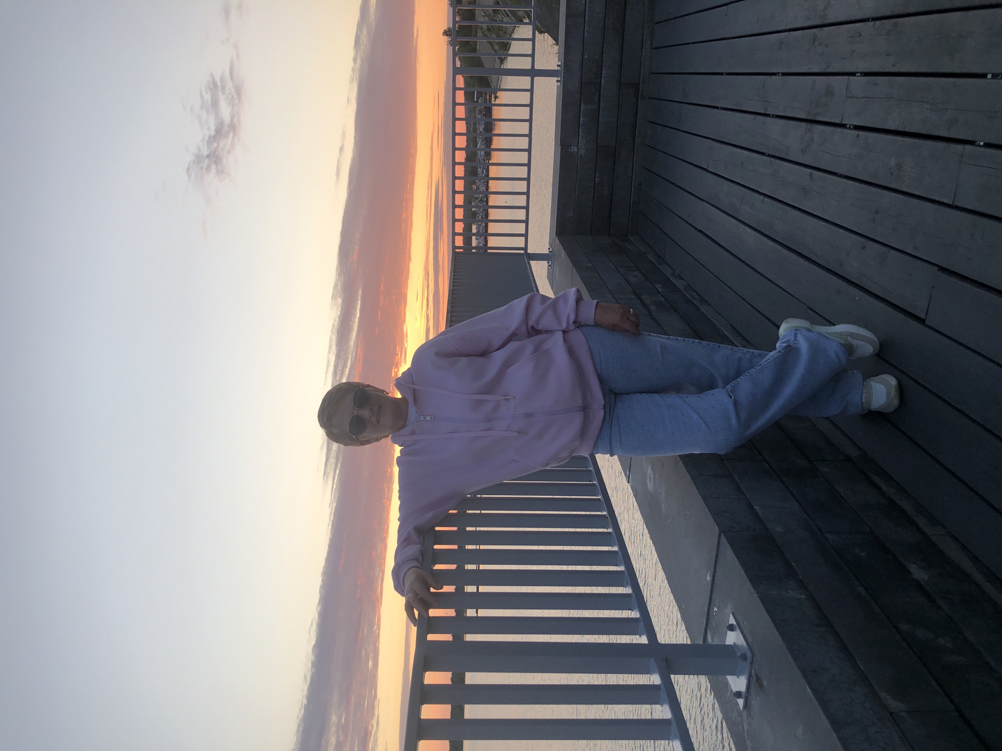 A woman stands on a wooden deck next to a metal railing overlooking a body of water at sunset. The sky is filled with orange and pink clouds. She is wearing a pink hooded sweatshirt, blue jeans, and white sneakers. She has short blonde hair and is wearing sunglasses. The location is believed to be in the city of Solikamsk, Russia.