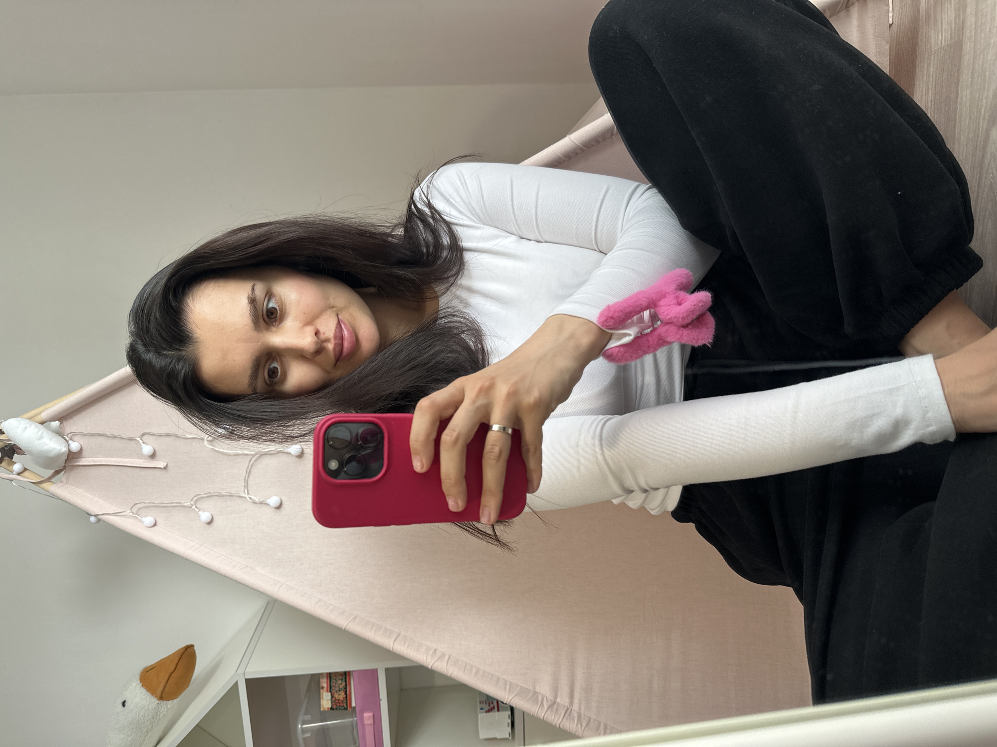 A young woman with long, dark hair, wearing a white long-sleeved top and black sweatpants, takes a selfie indoors. She is positioned inside or beside a light pink tent-like structure, possibly a teepee, which is adorned with a string of white, round fairy lights. Her right hand holds a bright pink-cased smartphone, with which she is capturing the image. A silver ring is visible on her left ring finger, and she has a pink furry accessory on her left arm.
Her expression is a slight smile, with her brown eyes wide open and looking directly at the camera. The background reveals a light-colored wall and some white shelving. On one of the shelves, a white stuffed animal with an orange beak, possibly a goose or duck, is visible. The lighting is bright and diffused, suggesting it is daytime. The overall setting appears to be a cozy, domestic space, possibly a child's room or a relaxed living area, located in Chaikovsky, Russia. No visible text is present in the image.