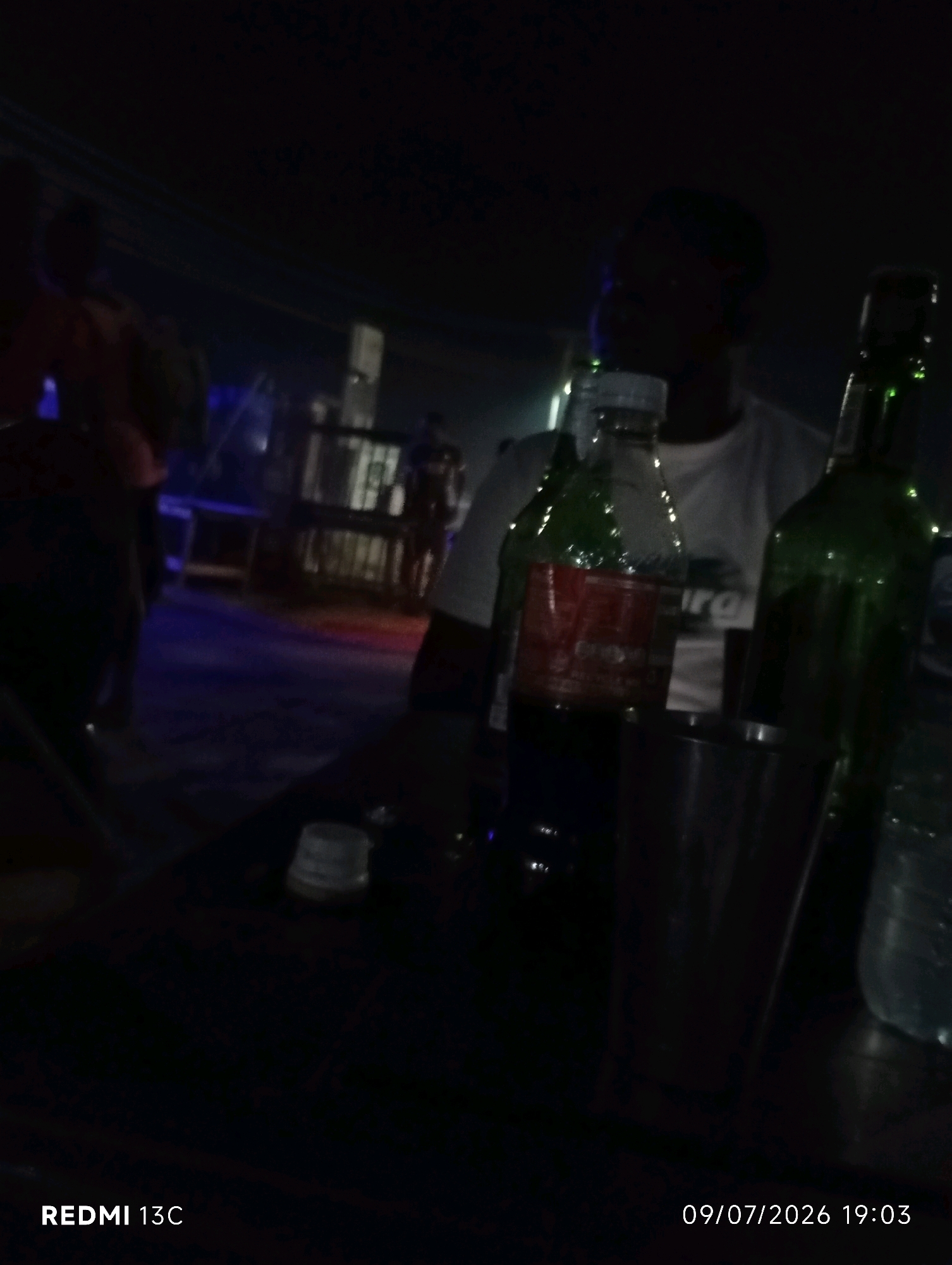 The image depicts a dimly lit outdoor setting at night, possibly a bar or gathering area. In the foreground, there are several bottles and a metal cup on a dark surface. One bottle is green and appears to be a beverage bottle, possibly beer or soda. Another bottle is red and has a label that is difficult to decipher due to the low light conditions. A metal cup is positioned between these bottles. A person is partially visible in the background, their face obscured by shadow. The overall atmosphere suggests a casual social gathering at night. The timestamp at the bottom of the image indicates the photo was taken on 09/07/2026 at 19:03. The "REDMI 13C" watermark suggests the image was captured using a Redmi 13C smartphone. The specific location in Shendam, Nigeria is not explicitly visible or inferable from the image content.