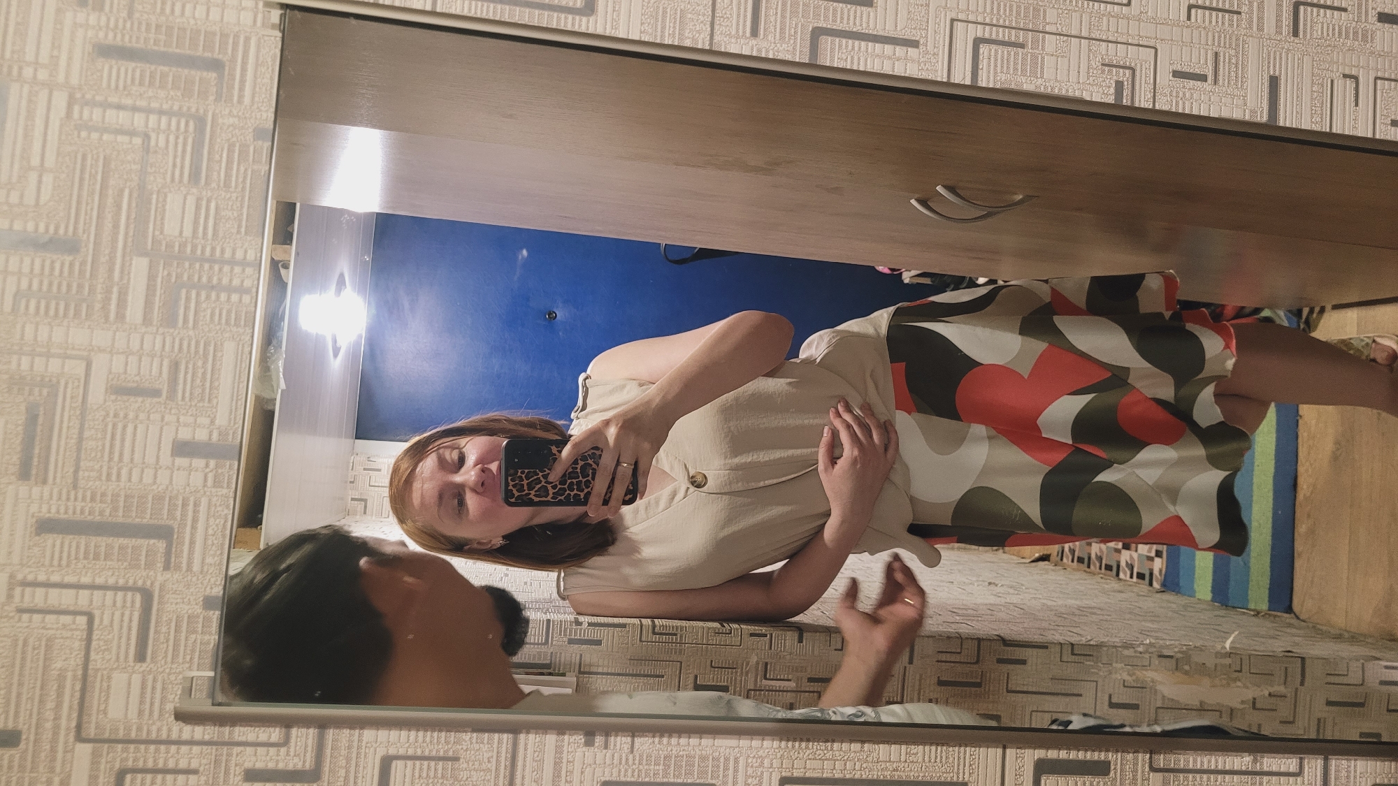 A man's head and shoulders are reflected in a mirror on the left, with his left hand reaching towards the right. In the center, a woman with auburn hair is reflected in the mirror, holding a smartphone with a leopard-print case to take a selfie. She is wearing a beige sleeveless top and a patterned skirt with red, white, olive green, and black shapes. Her right hand is placed on her stomach, and a ring is visible on her left ring finger. To the right of the woman's reflection, a portion of a wooden wardrobe is visible. The background of the reflection shows a blue door and patterned wallpaper. The foreground of the image shows wallpaper on the left and a wooden floor with a blue and green striped rug leading towards the wardrobe. The scene appears to be indoors, possibly in a hallway or bedroom.