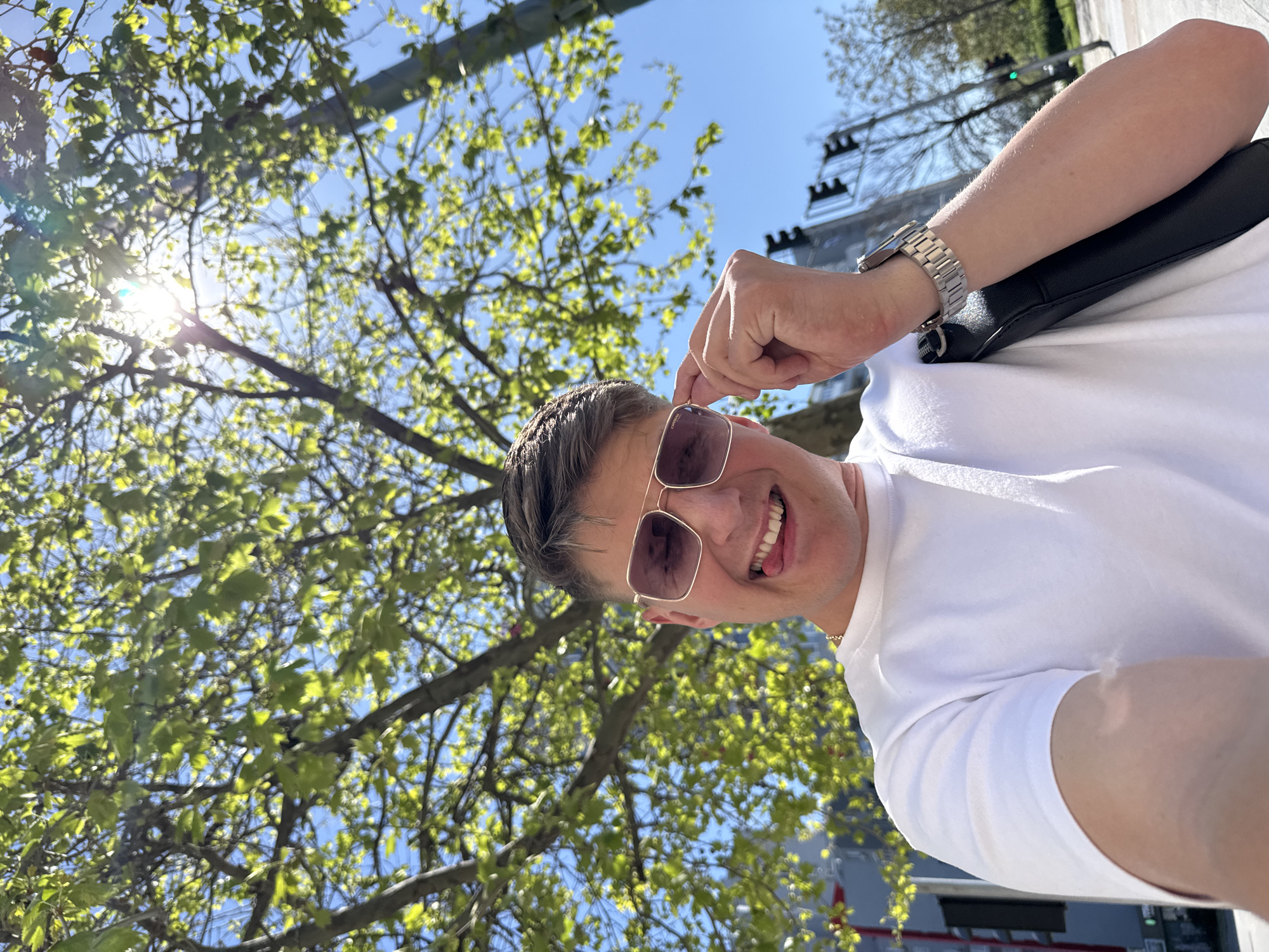 A medium shot captures a smiling man outdoors, tilting his head while holding his sunglasses. The man is wearing a short-sleeved white shirt, a wristwatch, and a black bag is slung over his shoulder. A tree with light green leaves is in the background, partly obscuring the blue sky. A glimpse of a building and a traffic light suggest an urban setting. The scene is likely in Plovdiv, Bulgaria, but this cannot be verified from the visual context.