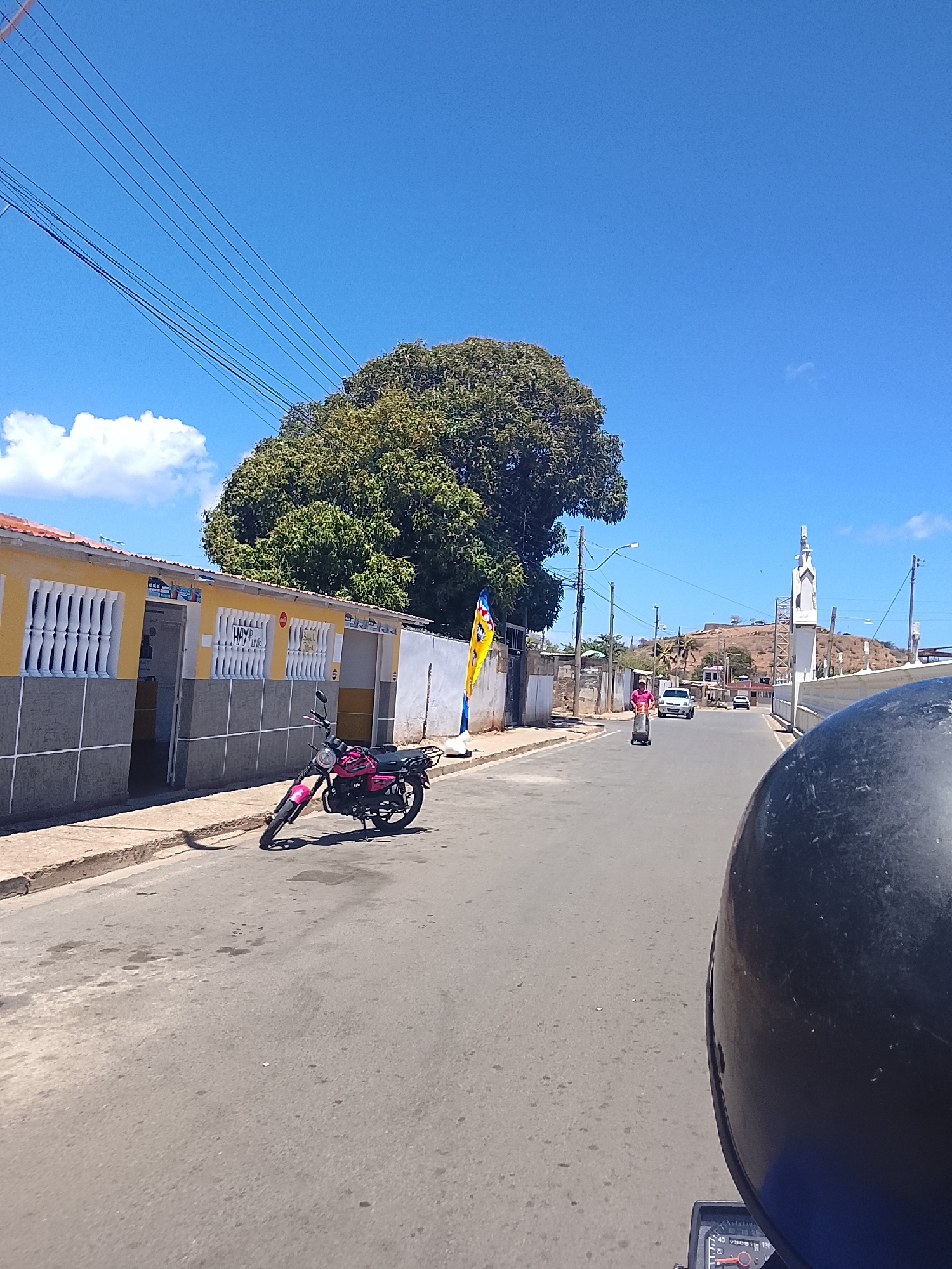 This media file presents a street-level view from the perspective of someone on a motorcycle, looking ahead on a bright, sunny day. The scene depicts a quiet street in what appears to be a small town or semi-rural area, consistent with Juan Griego, Venezuela.
In the immediate foreground, the top of a black motorcycle helmet and a small section of a motorcycle's dashboard are visible on the right, framing the shot.
On the left side of the road, a pink motorcycle is parked on the street next to a yellow building. This building features white decorative grilles over its windows and a gray base. Partially visible text on the building reads "HAY" and "VINE." Behind the building, a large, leafy green tree dominates the left midground, its foliage dense under the bright sun.
The paved road extends into the distance, gently sloping uphill. A person in a pink shirt is visible walking in the midground, pushing a two-wheeled cart or dolly. Further up the road, a white car and another vehicle are present. Utility poles with multiple wires traverse the upper left portion of the image against a clear blue sky.
On the right side of the road, further down, stands a prominent white structure with a tall, pointed element, resembling a church or a religious shrine, situated on the approach to a natural hill. The hill rises in the background, featuring some scattered vegetation and faint signs of development. The sky is largely clear blue with only a couple of small, wispy white clouds. The strong sunlight indicates midday conditions.