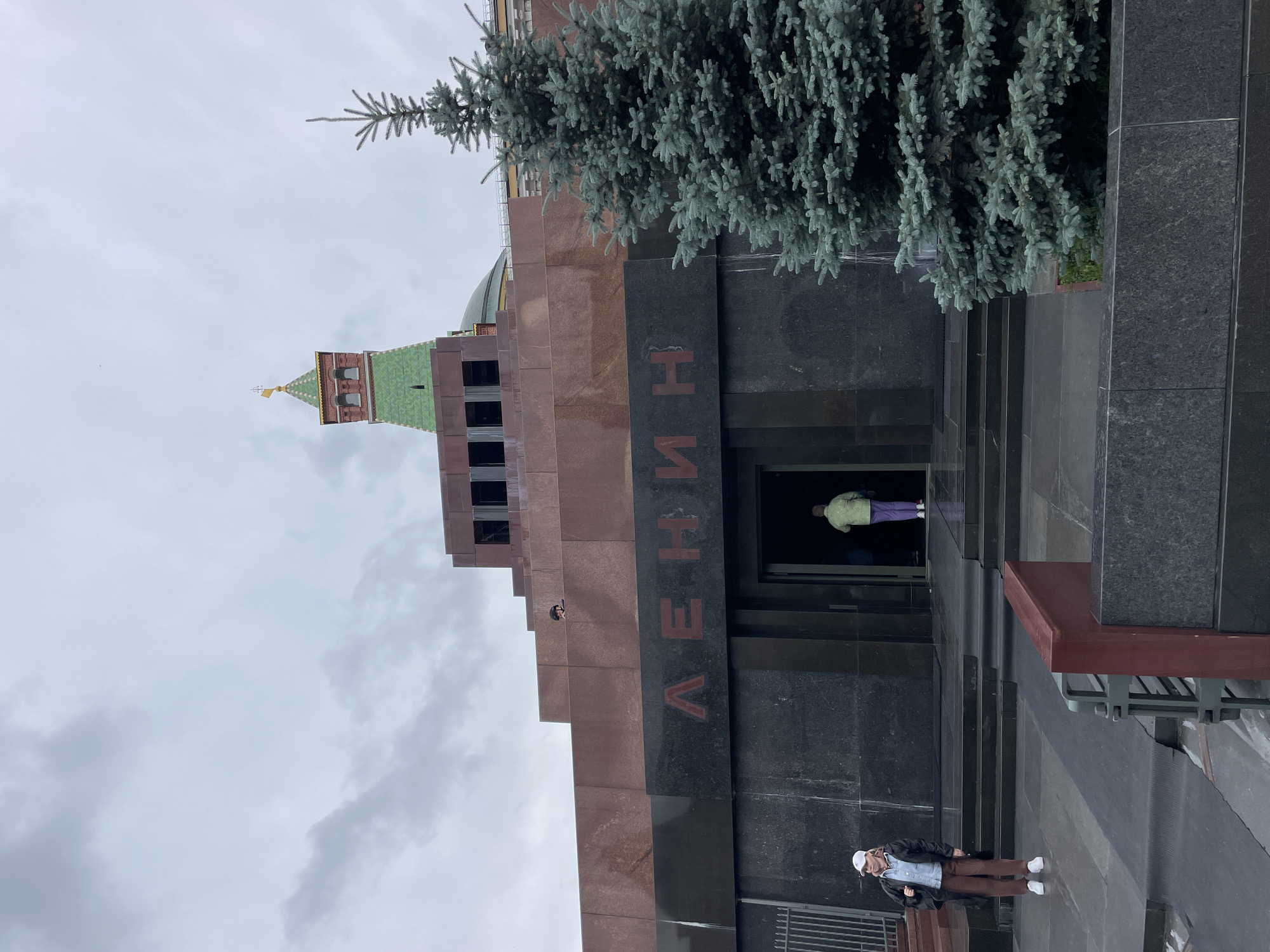 The image captures a view of Lenin's Mausoleum in Moscow, Russia, under a cloudy sky. The mausoleum, constructed from granite and marble, features a stepped pyramid design. The name "ЛЕНИН" (Lenin) is prominently displayed in large red letters on the dark granite facade.
In the foreground, a young woman wearing a white cap, a denim shirt, a black jacket, and brown pants stands on the left, facing slightly away from the mausoleum. To the right of the entrance, a tall, blue-green spruce tree is visible.
Two individuals are entering the mausoleum through an open doorway. One person, wearing a light green jacket and purple pants, is walking through the entrance. The cloudy sky and the somber architecture of the mausoleum create a somewhat solemn atmosphere. The overall scene depicts a public space with historical significance.