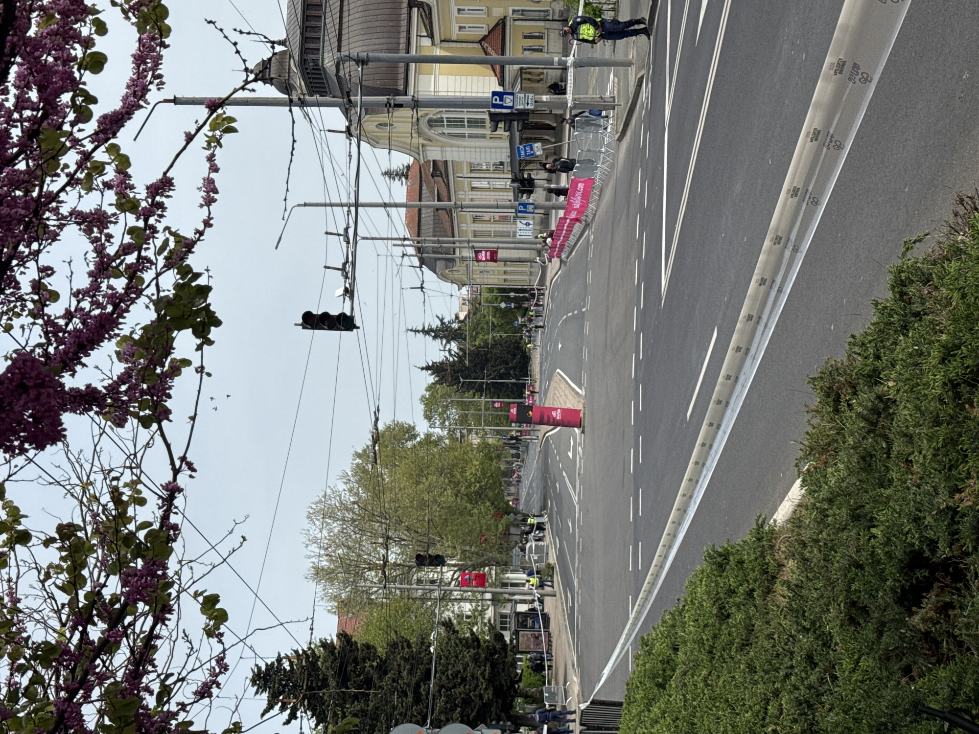 The image shows a street in Burgas, Bulgaria, with a closed-off section for a running event. A long stretch of asphalt road with white lane markings is visible. Barriers line the side of the road, some adorned with advertising. Traffic lights hang above the street. Buildings are present in the background. Trees are present on the left and right, with foliage and some blooming with purple flowers on the left. Several people, including police officers in high-visibility vests, can be seen along the barriers.