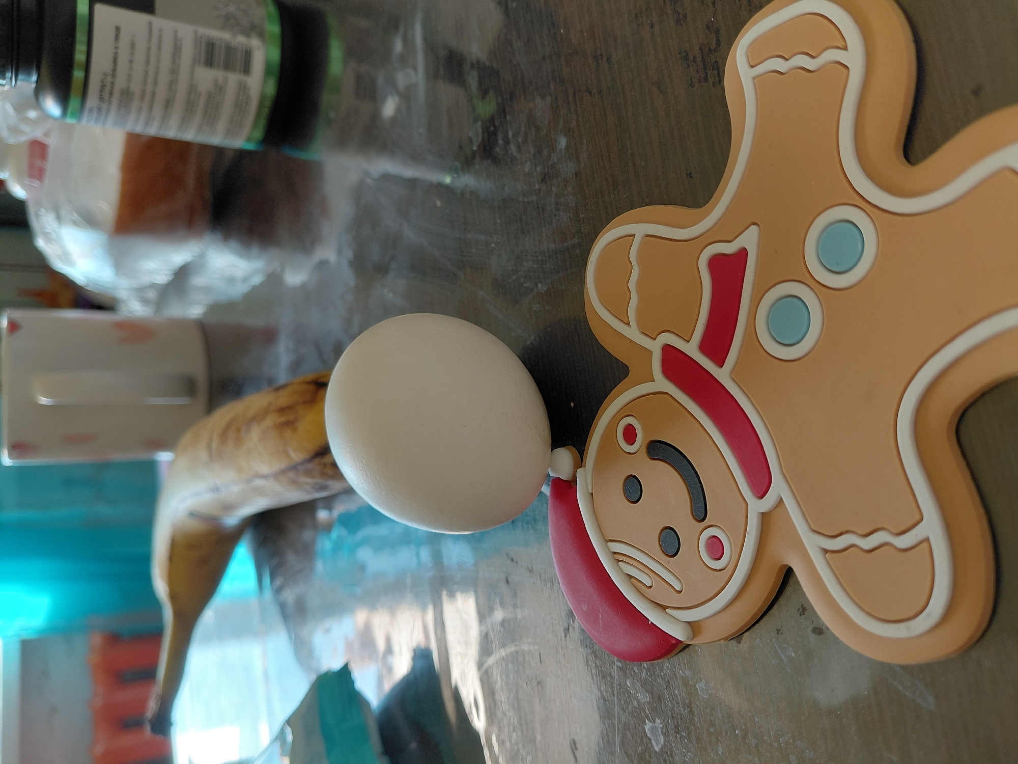 The image captures a peculiar still life composition on a dark, reflective surface, likely a table or countertop. A white, whole egg is prominently featured, balanced upright on the head of a gingerbread man-shaped silicone mat or coaster. This gingerbread figure is light brown with white outlines, a sad or grumpy expression, and wears a red Santa hat or hood, with light blue circular details on its body.
To the left of the egg, a ripe yellow banana with brown spots rests on the surface. The background is partially out of focus, revealing several other household items. A dark bottle with a green label, possibly a supplement container, is visible, showing faint Cyrillic text (e.g., "ИП" or "ИТРА") and a barcode. A white mug or cup can be seen behind the banana, and further back, a white plastic bag contains a brown or orange food item, possibly bread or cake.
The scene takes place indoors, likely in a kitchen or dining area. No specific activity is depicted beyond the arrangement of objects. The reflections on the surface suggest a glossy material.