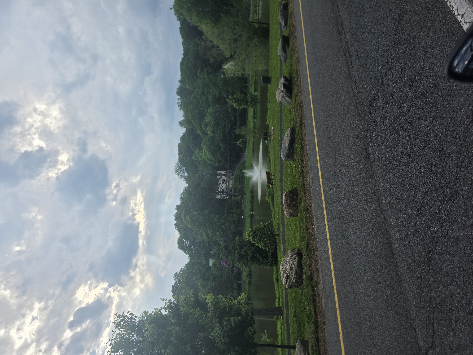 The image shows a park scene in New Rochelle, United States. A body of water, possibly a pond or lake, is in the center of the scene, featuring a fountain spraying water upwards. The water is surrounded by lush greenery and trees. A white building is visible in the background. A paved road with a yellow line is in the foreground. Large rocks are placed sporadically between the road and the park's grassy area. The sky is filled with clouds. A side mirror of a car is visible in the top right corner of the image.