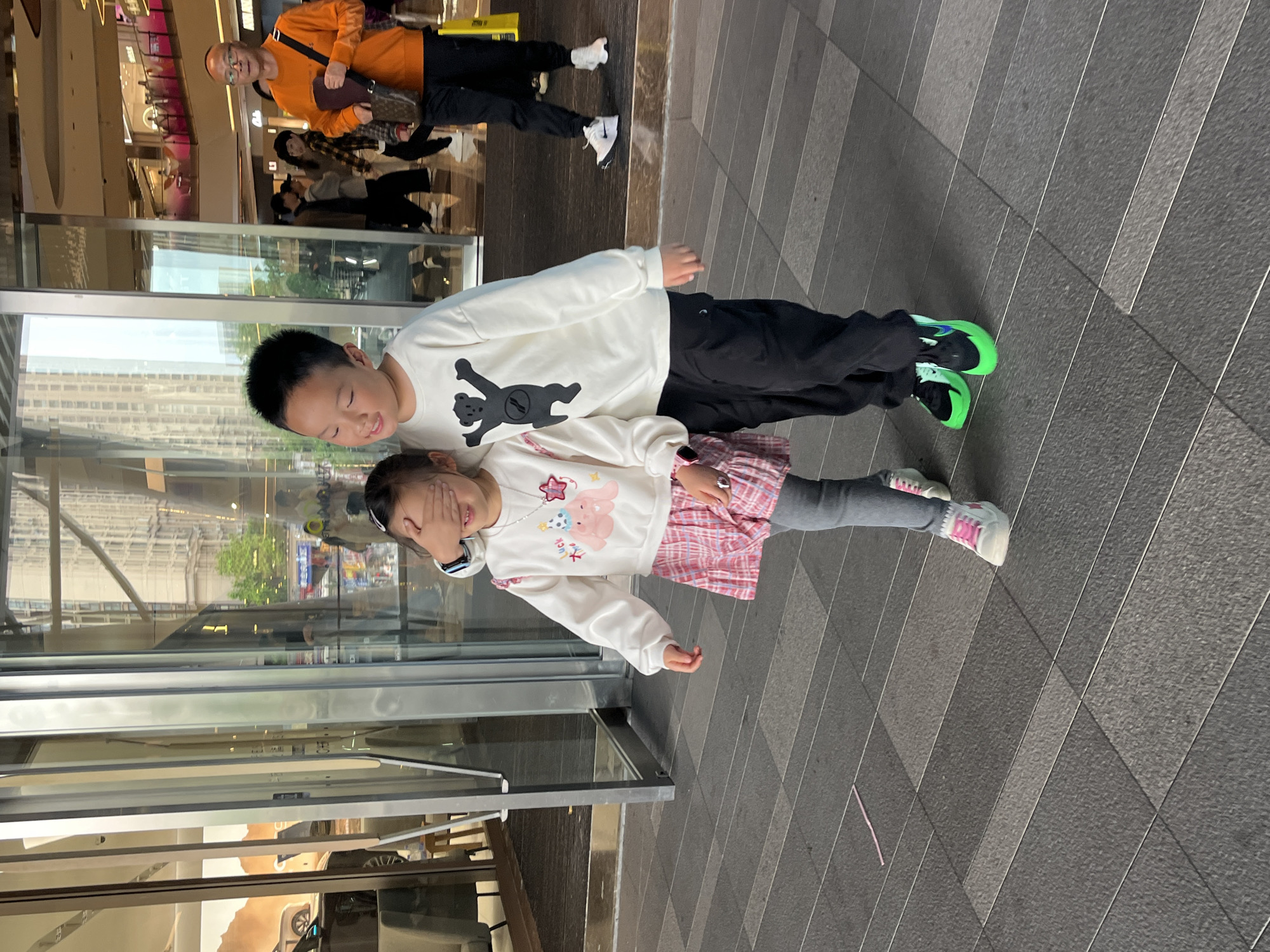 Two children are lying horizontally on a paved surface next to a large glass window. The older child, a boy, is on top, wearing a white long-sleeved shirt with a bear design and black pants, and bright green sneakers. The younger child, a girl, is below him, wearing a white long-sleeved shirt with a pink design, a pink and white plaid skirt, and grey leggings with pink and white sneakers. She has her hand over her eyes. The reflection in the glass suggests a cityscape. In the background, an older man wearing an orange shirt and carrying a brown bag is walking away. Other people are visible further back, inside what appears to be a shopping mall. The location is Liangjiang, China.