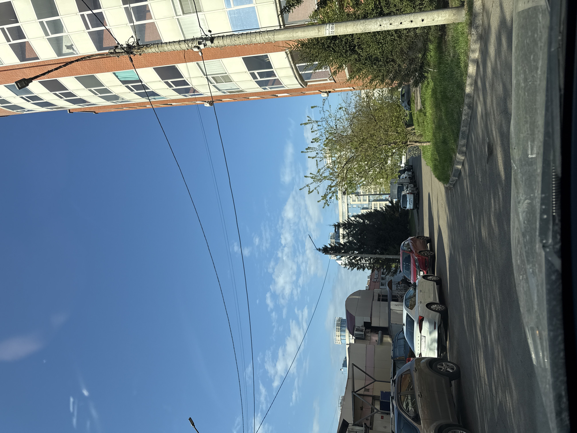 The image depicts a street scene in Krasnoyarsk, Russia. The view is from inside a vehicle, likely a car, looking out onto a street with parked cars on the right side. The cars include a tan SUV, a white sedan, and a red car. A sidewalk runs alongside the road, with a grassy area and trees separating it from a building further back. On the left side of the image, a tall residential building with a distinct grid-like pattern of windows is visible. Utility poles and wires crisscross the sky, which is a clear blue with scattered clouds. Several other buildings and structures are visible in the distance.