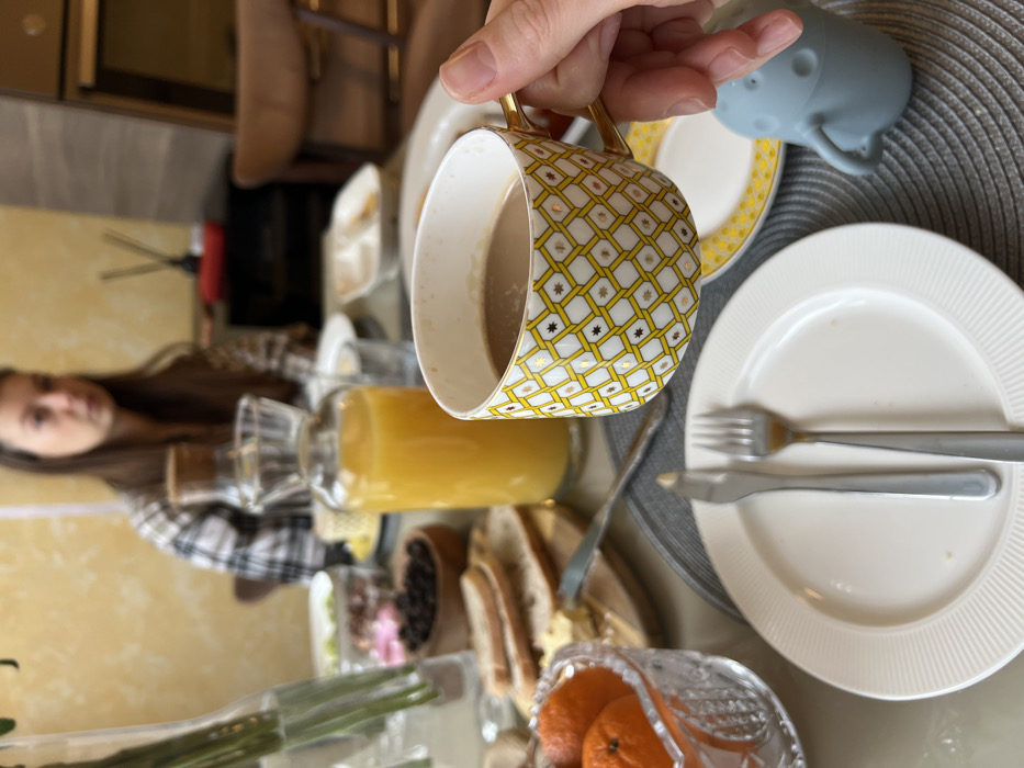 The image presents an indoor breakfast scene in Khimki, Russia. In the immediate foreground, a right hand holds a white teacup with a yellow and gold geometric pattern, containing a light brown liquid. Directly below the teacup, an empty white plate with ridged edges holds a silver knife and fork, resting on a dark grey woven placemat. To the right of the plate, a light blue silicone sippy cup or toy is partially visible. The table is set with various breakfast items, including a clear glass carafe filled with orange juice, a wooden cutting board with sliced bread and a pat of butter, a clear glass bowl containing whole mandarins, and a small wooden bowl holding dark raisins. Further back on the table, additional blurred bowls with various food items are discernible. A woman with long brown hair, wearing a plaid shirt, is seated at the table in the background, facing slightly right and looking away from the camera. The background features a textured yellow wall and blurred interior elements, including dark furniture and what appears to be a red-colored object, possibly a diffuser, on a distant surface.