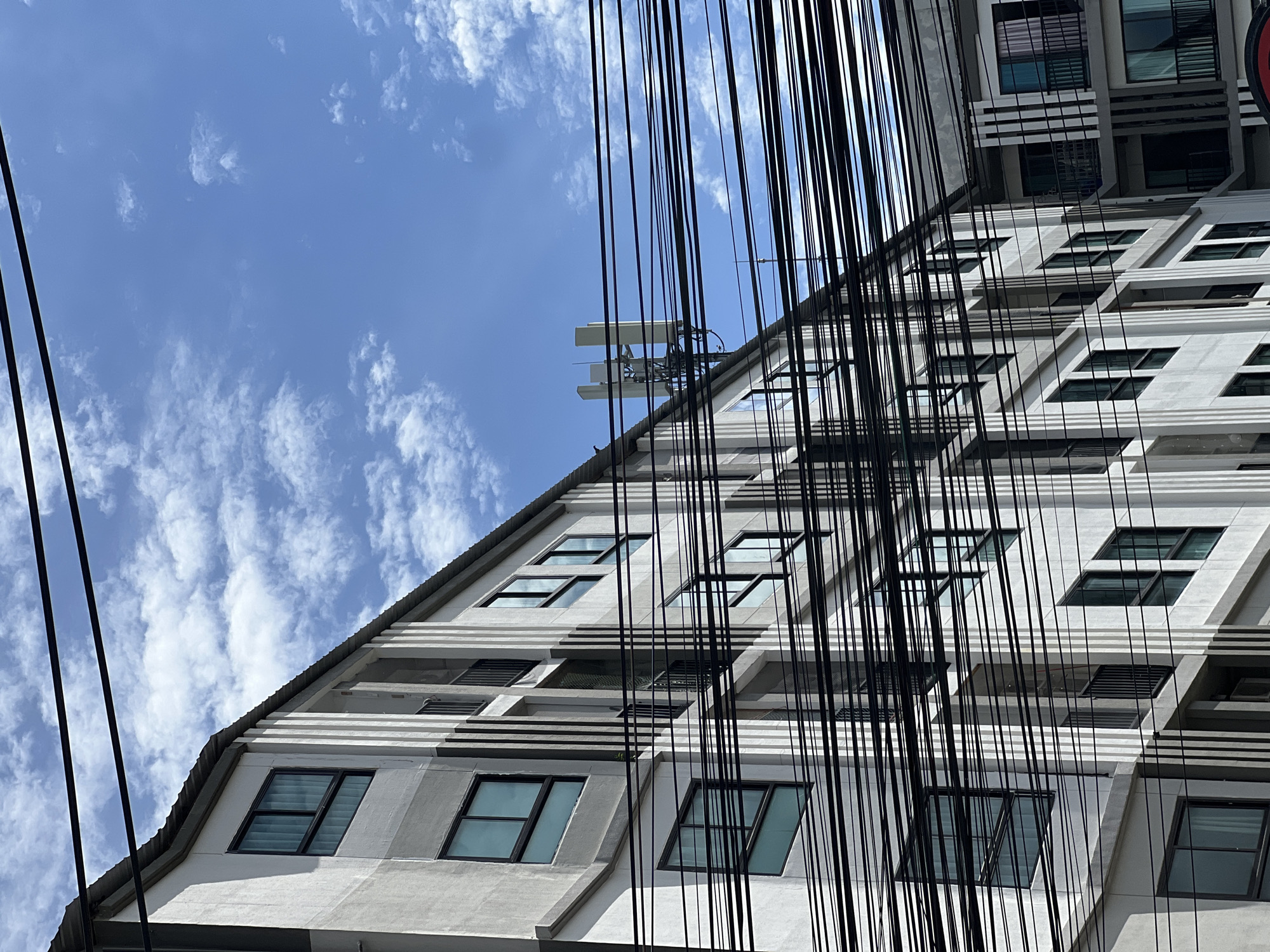 An outdoor, medium-shot photograph taken from a low angle shows the side of a modern, multi-story apartment building against a bright blue sky with scattered white clouds. The building's facade is primarily white and gray, with a repeating pattern of vertical lines and numerous windows. Black electrical wires crisscross the foreground, obscuring parts of the building. On the rooftop of the building, a cellular antenna array is visible, consisting of multiple panel antennas mounted on a metal structure. A single bird is perched on the edge of the roof, towards the left side of the frame.
