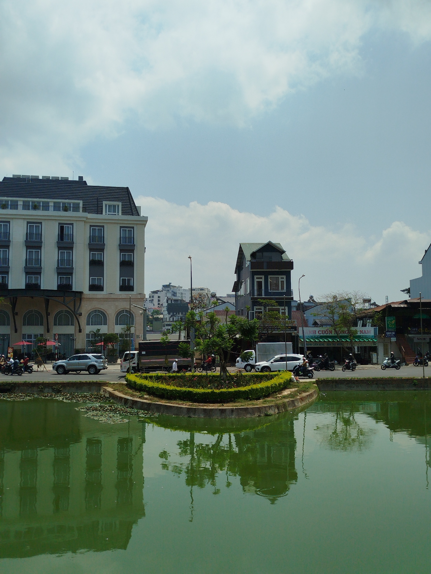 This photograph captures a cityscape in Da Lat, Vietnam. The foreground features a body of water with a green tint, reflecting the surrounding buildings and trees. An island-like structure sits in the middle of the water, adorned with green bushes. A road separates the water feature from the buildings.
On the left stands a tall building with a white facade, arched windows, and a dark roof. To the right is a building with a blue facade, partially obscured by trees. Street-level establishments line the road, with signage visible in Vietnamese. Cars, trucks, and motorbikes are on the street. A few people are standing near the water, and others are riding motorbikes. The sky above is a mix of light blue and white clouds.