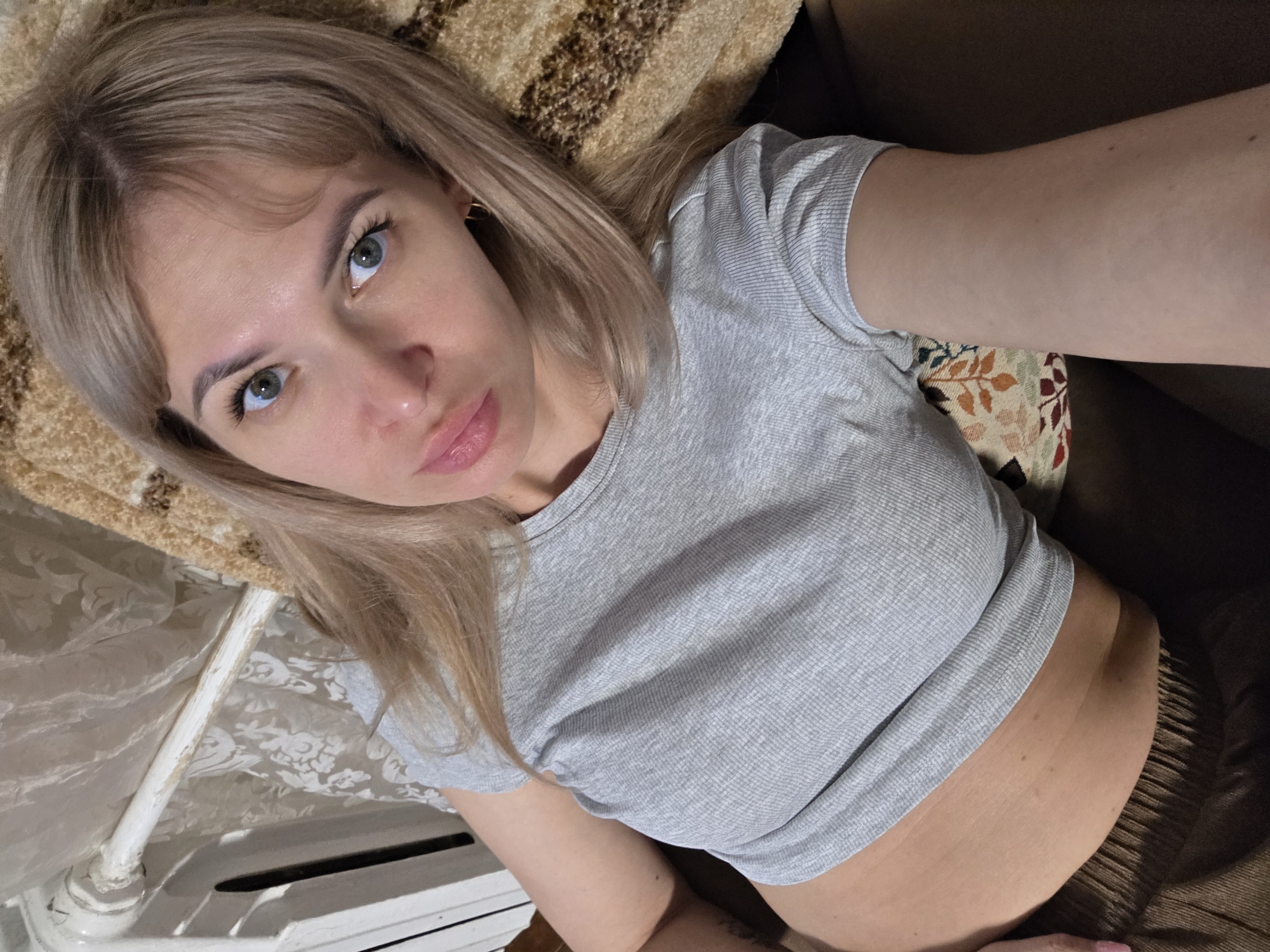 A woman with blonde hair and light eyes is lying down, wearing a gray t-shirt. She is indoors. A heating radiator is visible near her. The location is Chaikovsky, Russia.