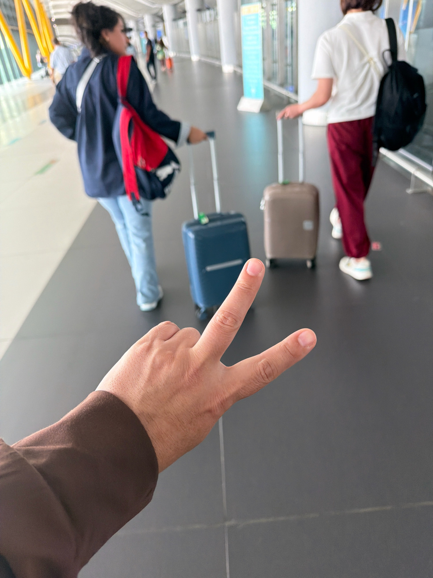 In an airport terminal in Arnavutköy, Turkey, a person's hand is in the foreground, displaying a peace sign. Behind them, two individuals are walking with luggage. The person on the left wears a dark blue jacket, light blue jeans, and a red backpack. The person on the right is dressed in a white top and maroon pants, carrying a black backpack. Two rolling suitcases, one blue and one tan, are visible between them. The terminal features a grey floor and white walls with illuminated signage.