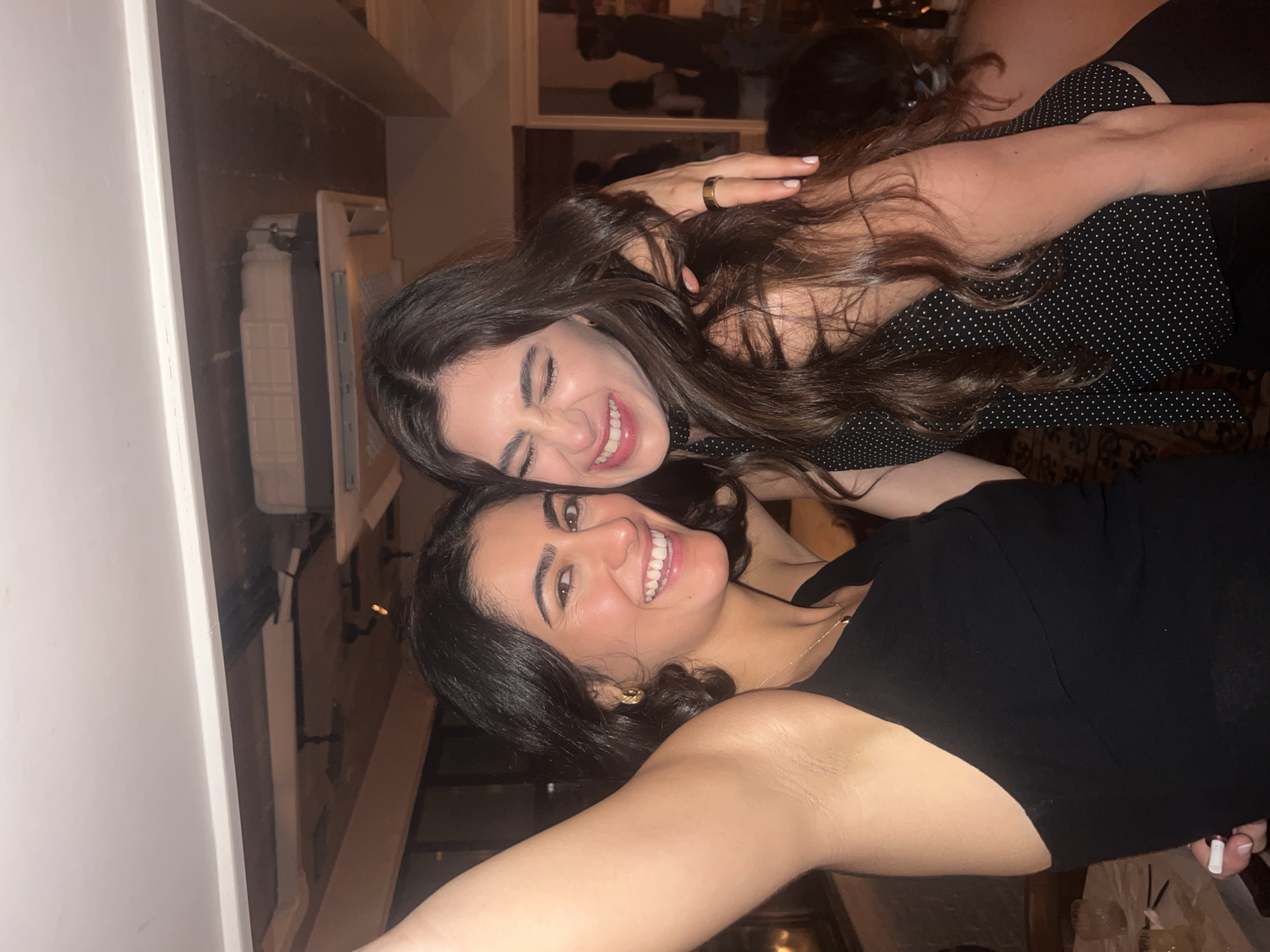 Two young women are posing for a photo indoors. The woman on top has long, wavy, dark brown hair, is wearing a black top with small white polka dots, and is winking. The woman below has dark hair, is wearing a black top, and is smiling broadly. The background is blurred, but hints of a mirror and a room setting are visible. The location cannot be determined to be Paris, France from the image.