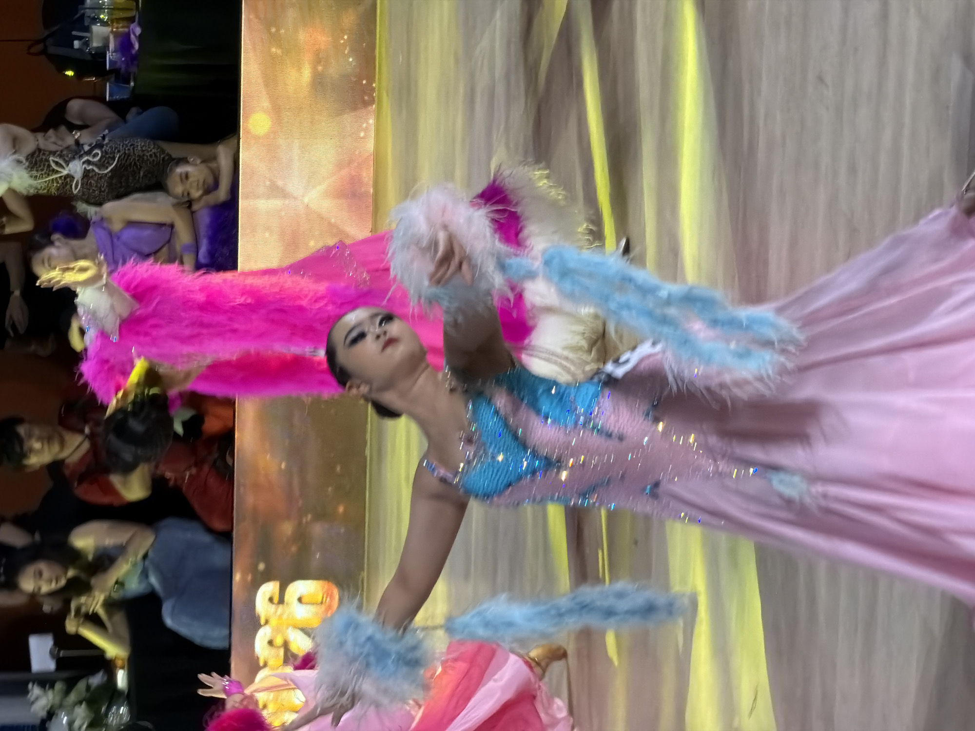 A young woman in a pink and blue sequined ballroom dance dress with feathered accents is captured in motion on a polished wooden floor. She is holding blue feathered props in her raised arms and looking to her left. The background is blurred, showing a large screen with orange and yellow glowing numbers and patterns. Several other individuals are visible in the background, some seated and others standing, all out of focus. The lighting is bright and theatrical, with yellow spotlights on the floor. The overall scene suggests a dance competition or performance event. There is no specific indication of the city of Hanoi, Vietnam.