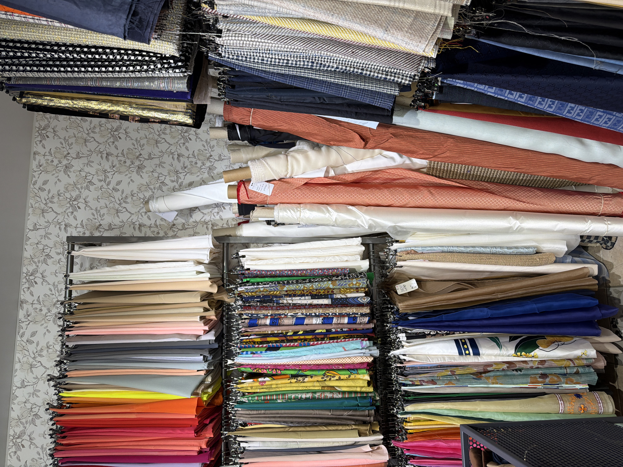 The image displays the interior of a fabric store or atelier in Saint Petersburg, Russia. The space is densely populated with textiles presented on metal racks and as upright rolls.
Three horizontal metal racks span the left side. The uppermost rack holds numerous solid-colored, lightweight fabrics, ranging from vibrant pinks, oranges, and yellows to greens, blues, greys, and finally pastels, beiges, and whites. The two lower racks display a diverse assortment of patterned and solid fabrics, including printed silks, scarves, and dress materials with floral, geometric, and abstract designs. A fabric piece with an embroidered border is partially visible on the bottom-left rack.
Central to the image, and extending rightwards, multiple fabric rolls stand vertically. These vary in size and texture, showcasing white, cream, peach, and terracotta hues, some with visible patterns or ribs. A tag is attached to one roll.
The right side features two vertical metal racks. The upper rack exhibits heavier, textured fabrics, including black, gold metallic, dark blue, a black and white geometric pattern, and various tweed-like materials in beige, grey, and yellow, some incorporating sequins or metallic threads. The lower rack displays additional suiting fabrics, including houndstooth, plaid, and a dark blue material with a repeating logo pattern.
The upper wall features light-colored wallpaper adorned with a subtle, repeating botanical pattern of leaves and blossoms in muted grey and beige. Illumination is consistent, and the floor is dark and largely obscured by the fabric displays.