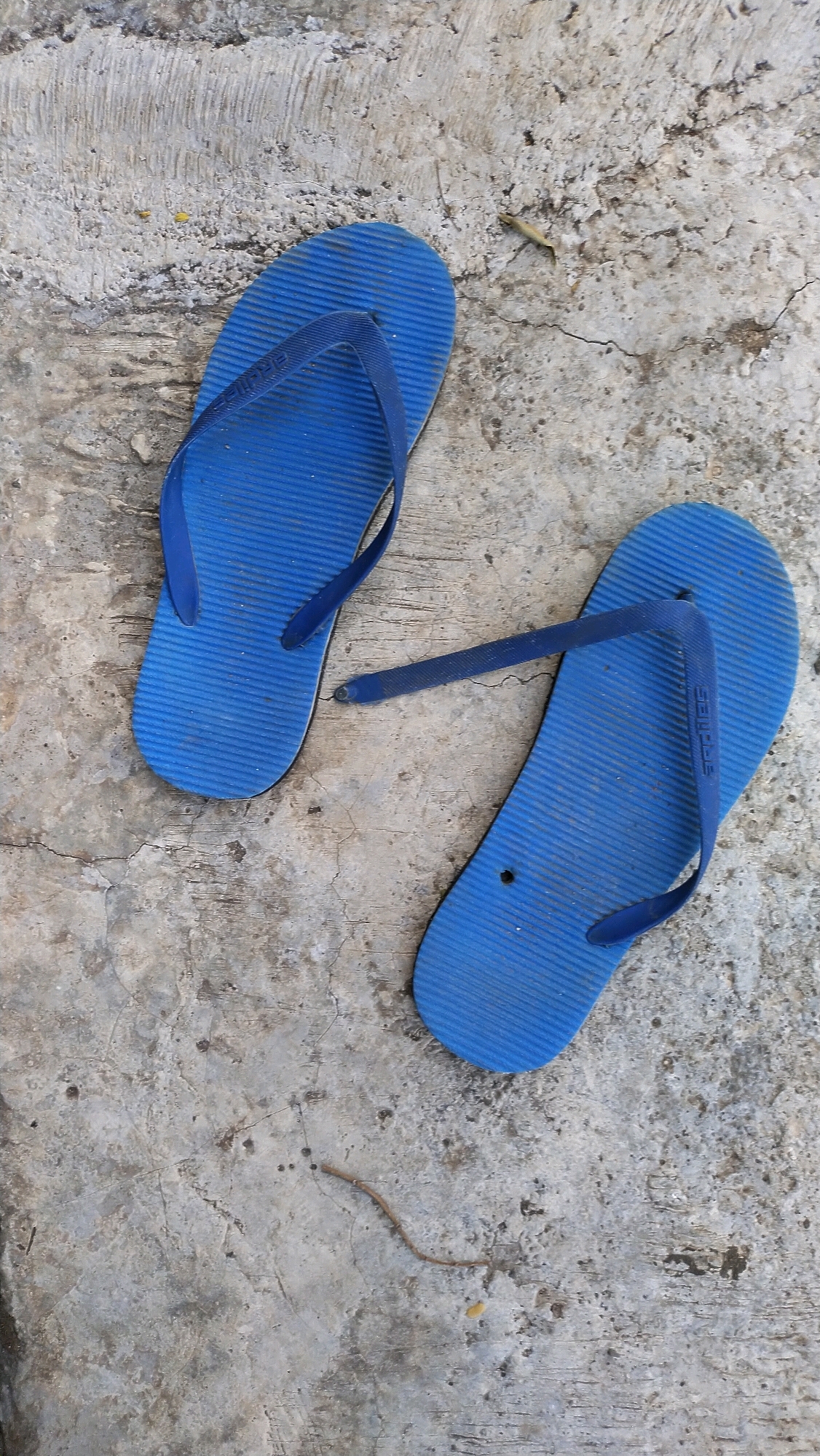 A pair of blue flip-flops lies on a cracked concrete surface. The flip-flops are positioned diagonally, with the left one higher and more to the left, and the right one lower and more to the right. The left flip-flop is complete, with its thong strap intact. The right flip-flop's thong strap is broken, with one part dangling and the other detached. The concrete surface shows signs of wear and tear, with visible cracks and textured markings. A small, dried twig is present on the concrete, to the right of the right flip-flop. The background is out of focus, but appears to be a continuation of the concrete surface. The image is taken from a top-down perspective. The location is the city of Klungkung, Indonesia.