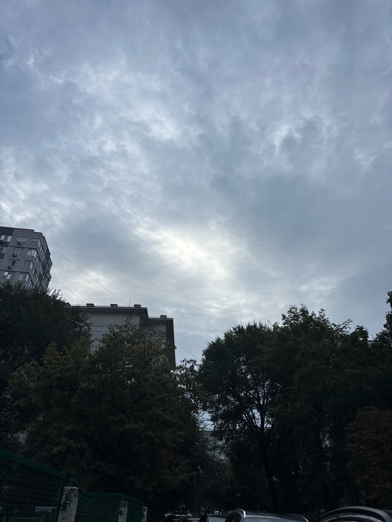 The image depicts a cloudy sky above apartment buildings and trees in Chișinău, Moldova. In the foreground, a dark green fence runs horizontally across the bottom of the frame. Behind the fence, dense foliage from trees obscures much of the lower part of the buildings and the ground. Two multi-story apartment buildings are visible, one on the left, taller and predominantly gray, and another on the right, with a lighter facade and visible rooftop details. The sky is filled with a variety of gray clouds, with a brighter patch suggesting the sun's presence behind them. Power lines are visible stretched between the buildings. The overall atmosphere is overcast and somewhat muted in color.
