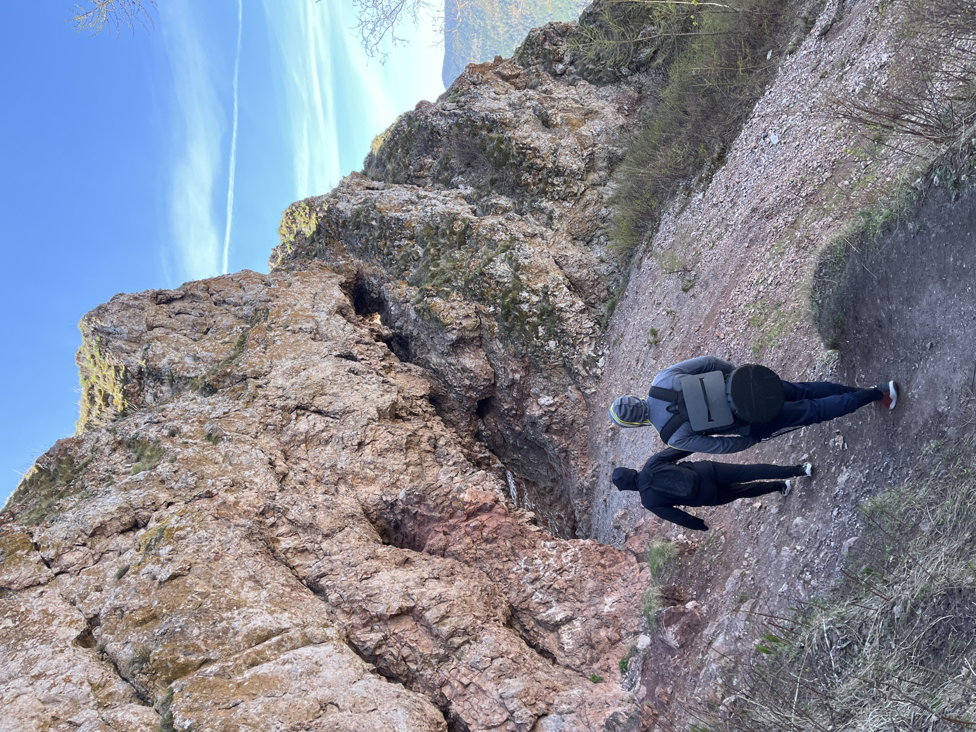 Two individuals are present in an outdoor setting, adjacent to a large rock formation. One individual is wearing a gray jacket, dark pants, and red shoes, and carrying a dark backpack. The second individual is wearing dark clothing. They are standing on a path next to the rock formation, which appears to have caves and crevices. The sky is blue with light clouds. The location is consistent with the Takmak rock climbing area of Stolby National Park in Krasnoyarsk, Russia.