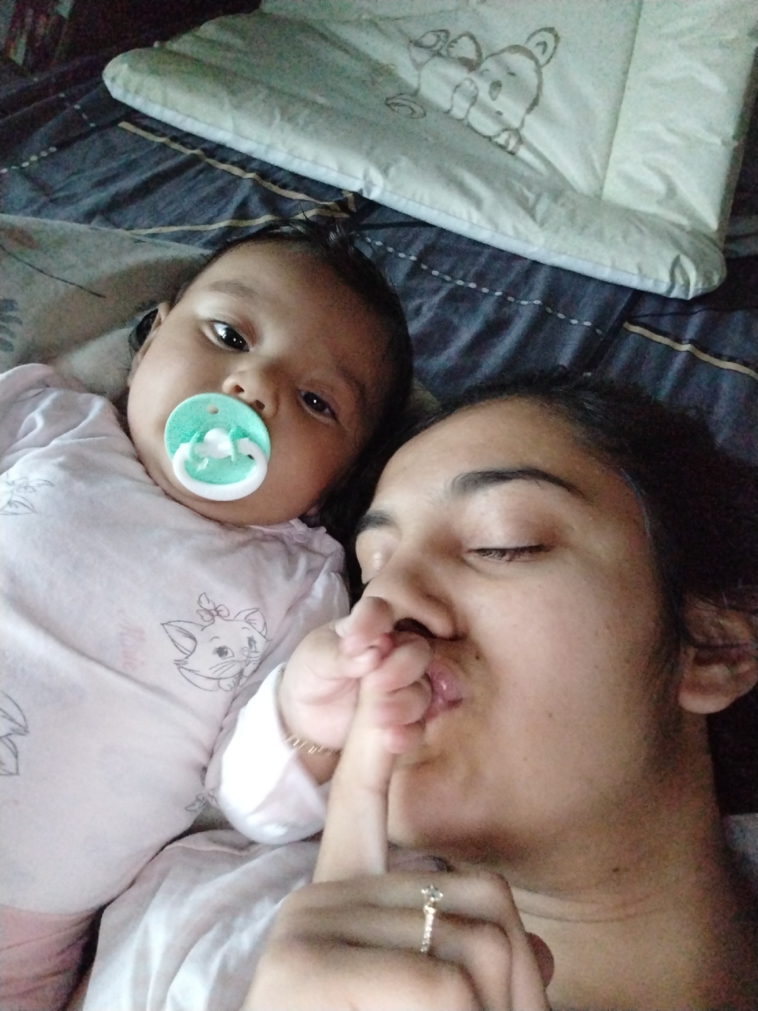 A photograph shows a woman and a baby lying on a bed. The baby is on the left with a pacifier in its mouth, wearing a light pink onesie adorned with a cat illustration. The woman is on the right, with her eyes closed and her finger placed over her lips in a 'shush' gesture. She is wearing a ring on her finger. The bed is covered with a blue and gray patterned sheet, and there is a pillow with a bear design in the upper part of the image. The location is unknown.