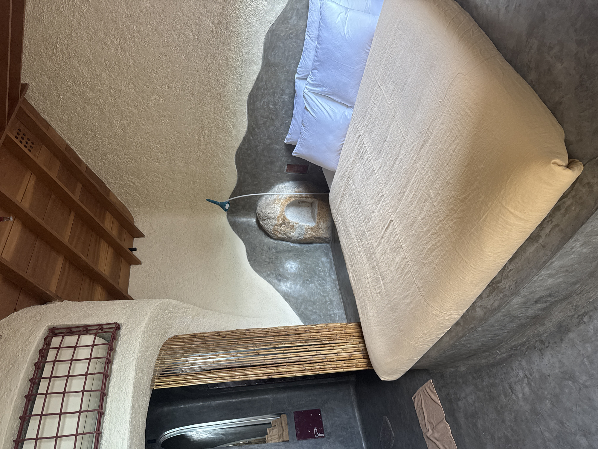 This image presents an interior view of a distinctively designed room, likely a bedroom or accommodation suite, characterized by its organic and rustic architecture. The perspective is from an elevated angle, looking down into the space.
A prominent feature is a large bed, centrally positioned and covered with a light beige or off-white textured duvet or blanket. Several fluffy white pillows are arranged at the head of the bed. The bed rests on an irregularly shaped, dark grey textured floor, which appears to be made of polished concrete or a similar material. This dark flooring contrasts with the light beige, textured walls and ceiling, which are sculpted with soft, rounded contours, giving the room a cave-like or adobe aesthetic.
Near the head of the bed, integrated into the floor's organic shape, is a unique, irregularly formed stone fixture, possibly a decorative element or an unconventional basin. A thin white cable runs near this stone, leading to a small, light blue object, which could be a soap dish or a small planter.
To the left side of the room, a multi-tiered wooden shelving unit is built into the wall, featuring small ventilation holes. Below the shelving, a window with a dark red metal grid is set into the contoured wall. Further down, beneath the bed, a horizontal bamboo screen or blind covers an opening, above what appears to be a dark-colored basin or tub. Adjacent to this, a small, dark rectangular sign with a white key icon is affixed to the wall. A small, light brown towel lies on the dark floor in the lower right corner.
The room's design emphasizes natural materials and curved forms, creating a unique and tranquil atmosphere. No people are present, and no specific activity is taking place. The setting, with its distinctive architecture and use of natural elements like wood, stone, and bamboo, aligns with the context of Bac Nha Trang Ward, Vietnam, suggesting a resort or eco-friendly accommodation.