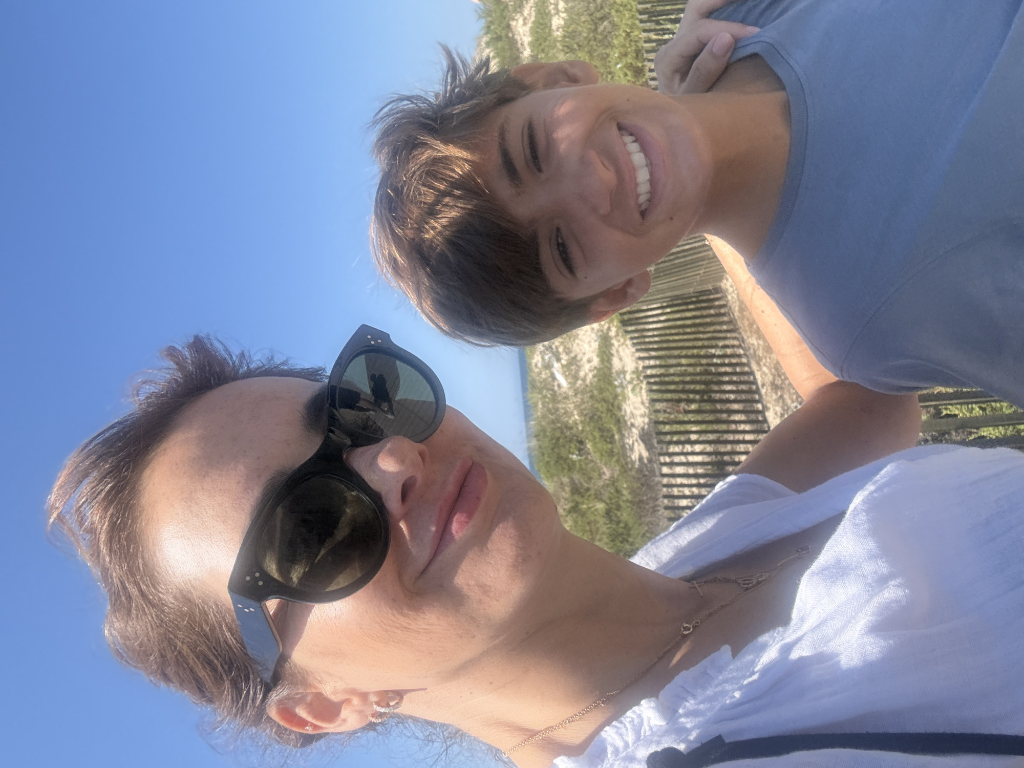A woman and a young boy are posing for a selfie on a sunny day, with a beach and ocean in the background. The woman, on the left, is wearing sunglasses and a white top. The boy, on the right, is wearing a blue t-shirt and smiling. They appear to be standing on a boardwalk or path, with a wooden fence and sand dunes visible. The location is identified as Vila Nova de Gaia, Portugal.