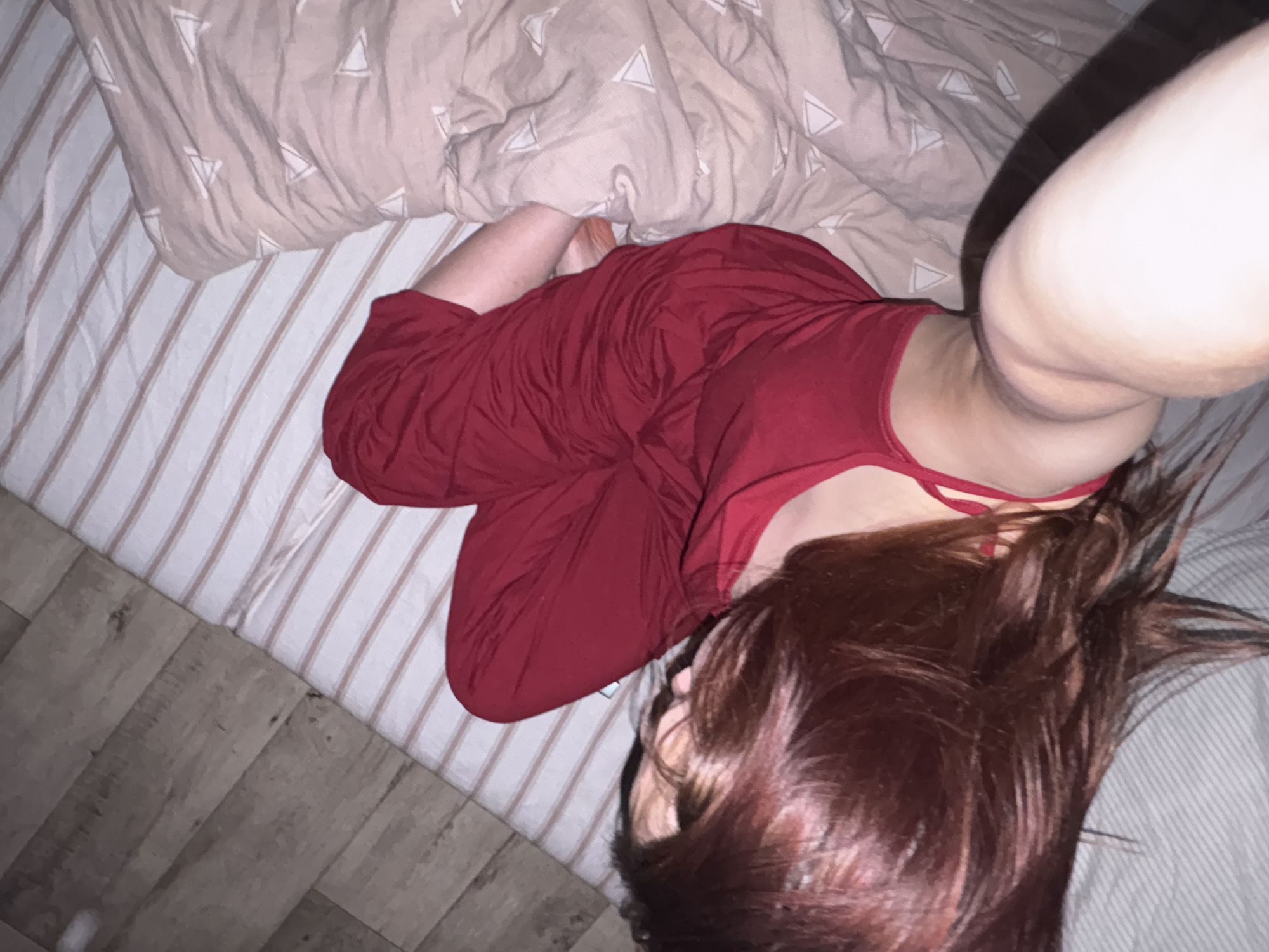 An overhead photograph captures a person lying on a bed in a domestic indoor setting, likely a bedroom, taking a selfie. The individual has reddish-brown hair spread across the pillow and is dressed in red clothing, consisting of a camisole-style top with thin straps and matching bottoms, possibly pajamas or lounge wear. Their right arm is extended, holding the camera, and is visible in the upper right portion of the frame.
The bed is adorned with white sheets featuring subtle, light brown vertical stripes. A beige duvet or blanket, patterned with scattered white triangles, is partially covering the person's lower body and is crumpled near the top of the bed. A segment of dark wooden or wood-patterned flooring is visible along the lower left edge of the image. The lighting appears dim, suggesting an evening or nighttime scene, or a softly lit room. No text is visible in the image. This scene is located in Прикамский, Russia.