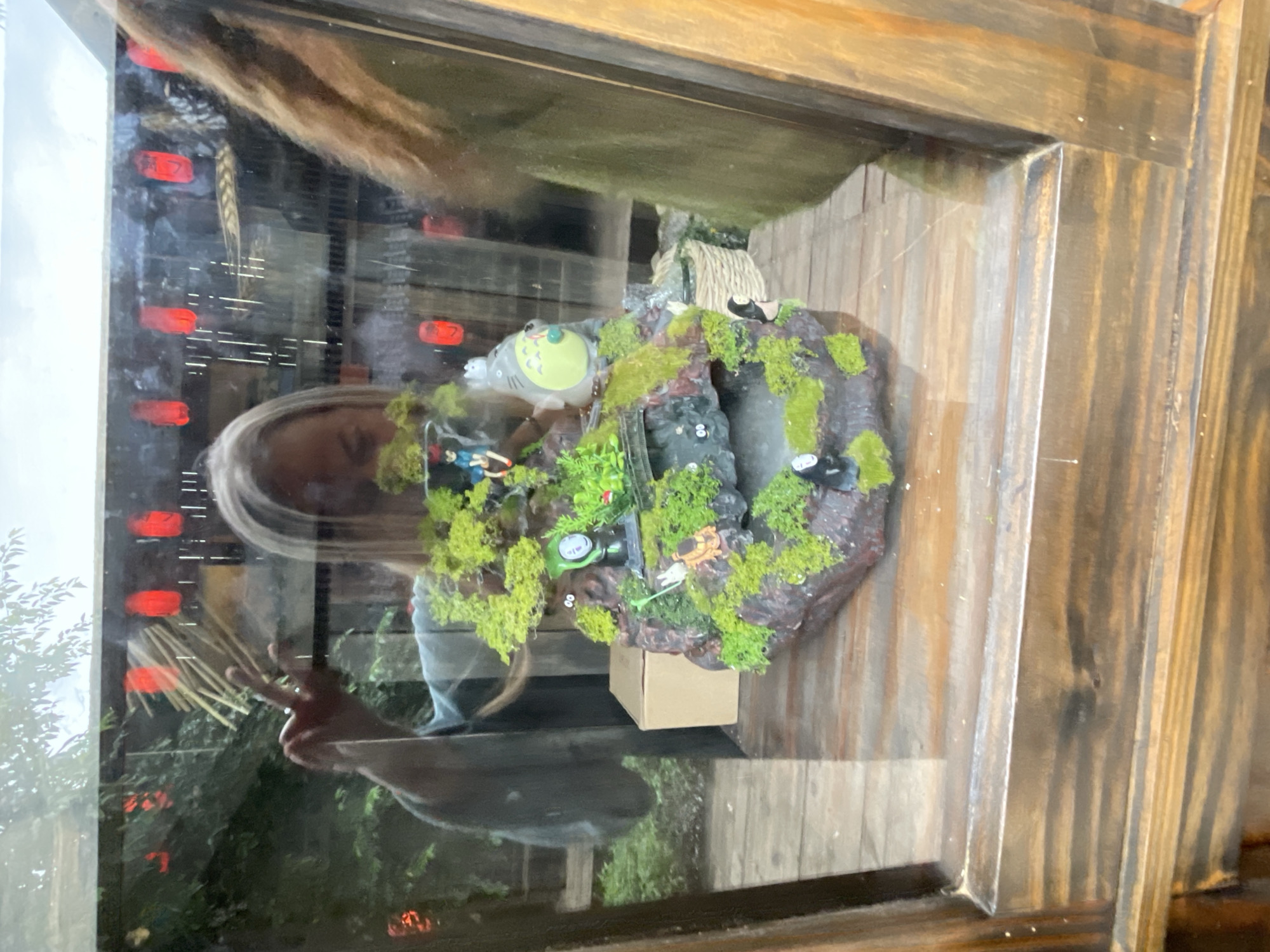 A close-up, angled shot captures a miniature diorama displayed behind glass. The diorama depicts a lush, green landscape with rocky outcrops covered in moss, miniature trees, and a small waterfall. Several small figures from the Studio Ghibli movie "Spirited Away" are visible, including Totoro, No-Face, and soot sprites. The diorama is situated on a wooden surface, and behind it, a cardboard box is visible. Reflections in the glass reveal a person with blonde hair, wearing a light-colored garment, making a peace sign with their fingers. Red lanterns hang in the background, and trees are visible through the glass. The overall scene appears to be an outdoor display, possibly in a garden or park area, given the presence of foliage and the style of the display. This scene is located at Cam Ly Waterfall, a popular tourist destination in Da Lat, Vietnam.