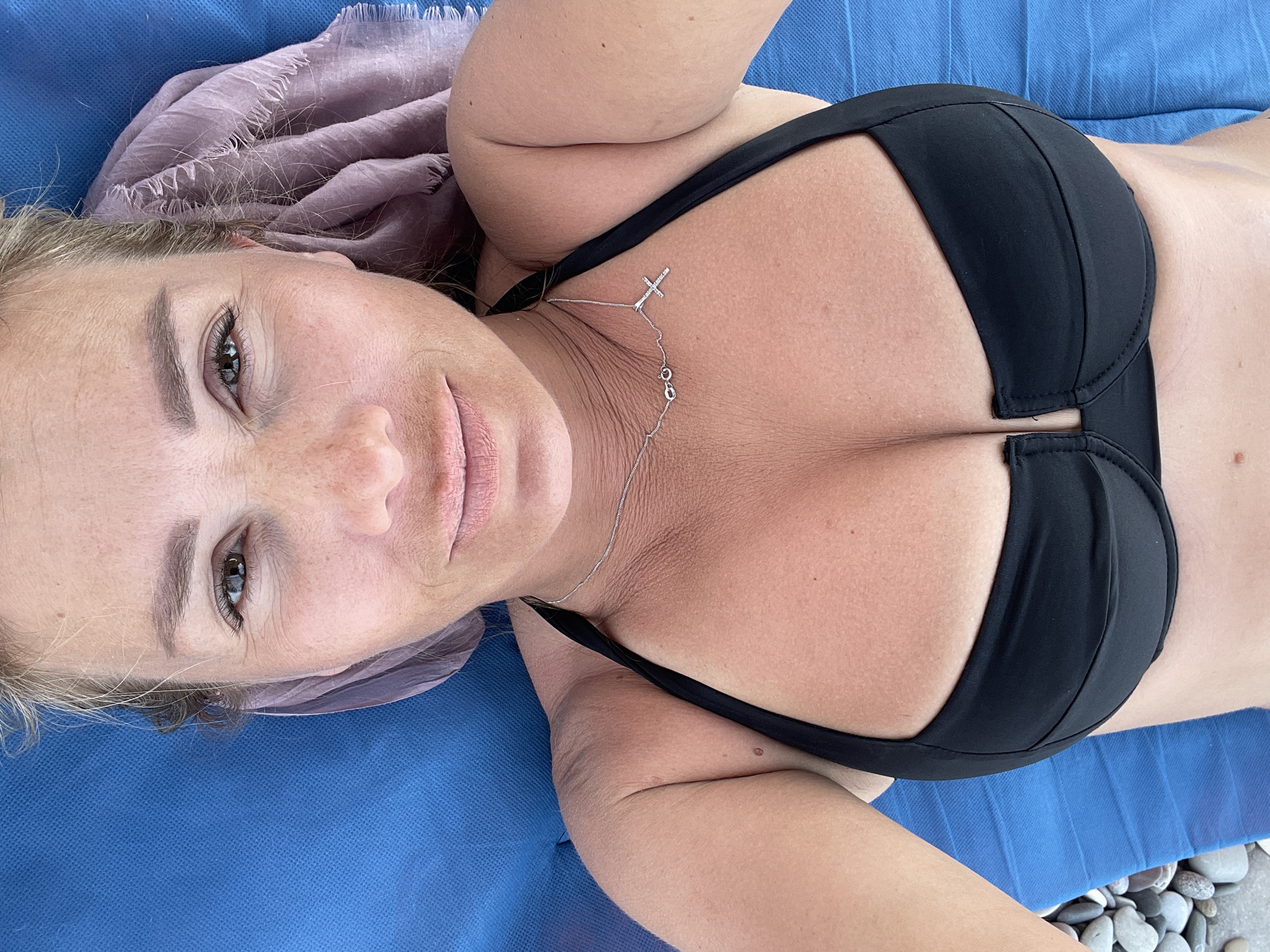 A woman is captured in a selfie taken from a high angle, lying on a blue lounge chair. She is wearing a black bikini top and a silver necklace with a cross pendant. Her hair is blonde and pulled back. A sheer purple scarf is draped behind her head. The background is out of focus, with hints of pebbles and the blue chair. The woman's skin appears tanned.