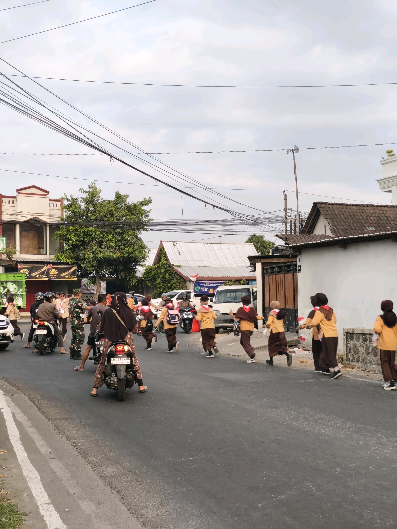 The image depicts a street scene in Desa Dibal, Indonesia, with a group of people, predominantly children in uniform, participating in an outdoor event. Several children, wearing brown and yellow uniforms with red neckerchiefs, are running or walking along the road, some carrying small flags. In the foreground, a person is seated on a motorcycle facing away from the camera, while another motorcycle is parked nearby. A person in military uniform stands on the road, seemingly directing or observing the group. Several other individuals, including adults on motorcycles and pedestrians, are scattered throughout the scene. The environment includes buildings with tiled roofs, trees, utility poles with numerous wires, and a cloudy sky. The road is asphalt with a white line marking its edge. The scene appears to be a daytime event, possibly a parade or a school-related activity, taking place on a moderately busy street.