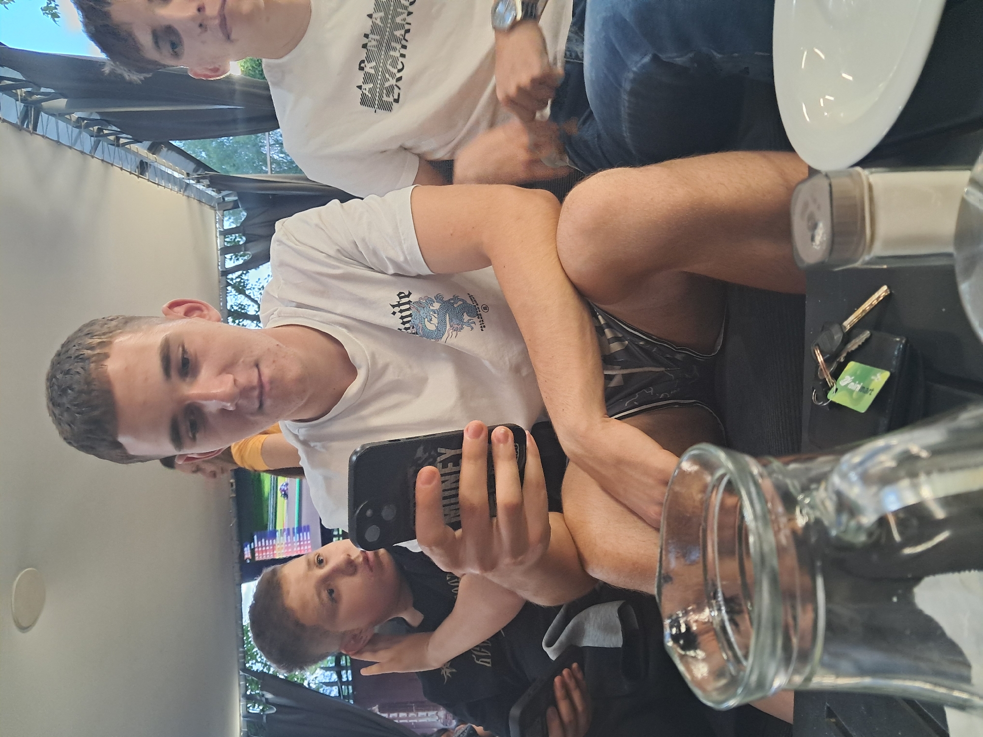 The image depicts a scene within a covered outdoor cafe or restaurant in the city of Blagoevgrad, Bulgaria.
Three young males and one partially visible female are present. A young male, positioned centrally, has short brown hair and is wearing a white t-shirt with a blue dragon graphic and the text "UNITE" and "FALL ALL THINGS RISE", along with dark patterned shorts. He is seated and looking down at a black smartphone held in his right hand. The phone case features the word "MONEY".
To his left, a younger male with light brown hair, wearing a black t-shirt, is seated. His right hand touches his ear, and his gaze is directed towards the central male's smartphone screen. A second smartphone is partially visible in the left male's left hand.
To the central male's right, another young male with light brown spiky hair, wearing a white t-shirt with "ARMANI EXCHANGE" printed on it and blue jeans, is seated. He wears a silver watch on his right wrist and is gazing to his right, not interacting with the smartphones.
In the background, a female wearing a yellow top is partially visible behind the central male's head. A large screen displays a racing video game or simulation, featuring a green track, a white car, and a digital scoreboard on the left. The environment includes a white ceiling, dark metal support structures, dark fabric canopies, and green foliage visible beyond the canopy structure.
In the foreground, a dark-colored table holds a clear glass pitcher filled with water, a set of keys with a green tag, a clear shaker with a brown top (likely a condiment shaker), and an empty white ceramic plate. The scene is illuminated by natural daylight.